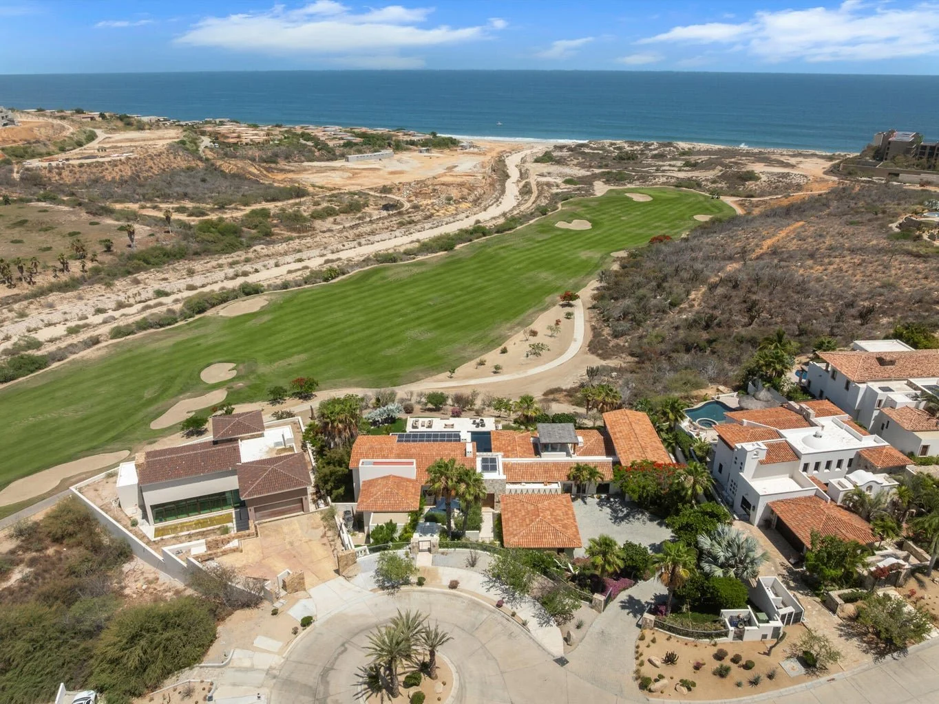 Aerial view of a residential neighborhood adjacent to a golf course with a coastline in the background. The area features modern white houses with red tile roofs, lush greenery, and a sandy terrain.