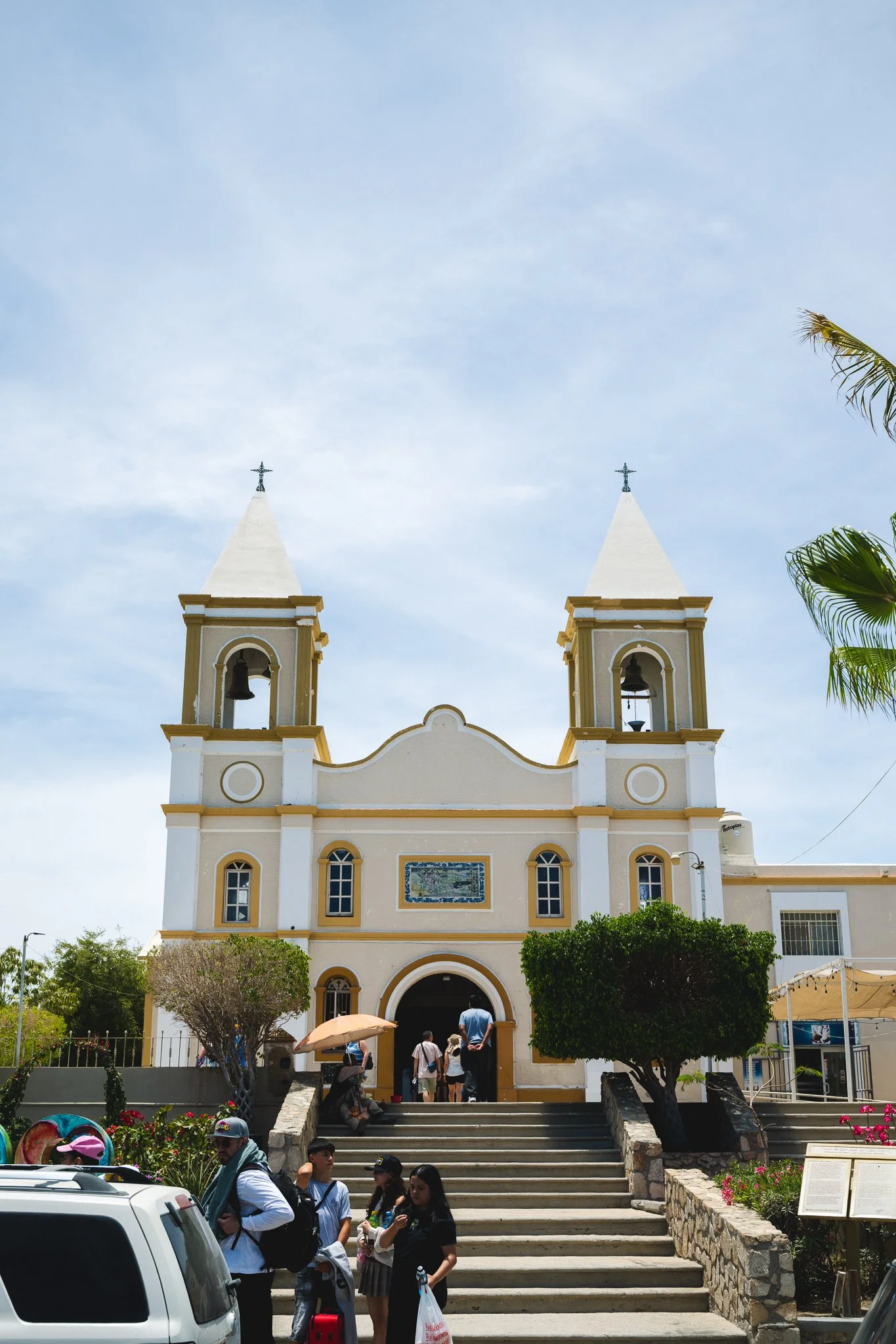 People waiting outside a church with tall twin steeples on a sunny day.
