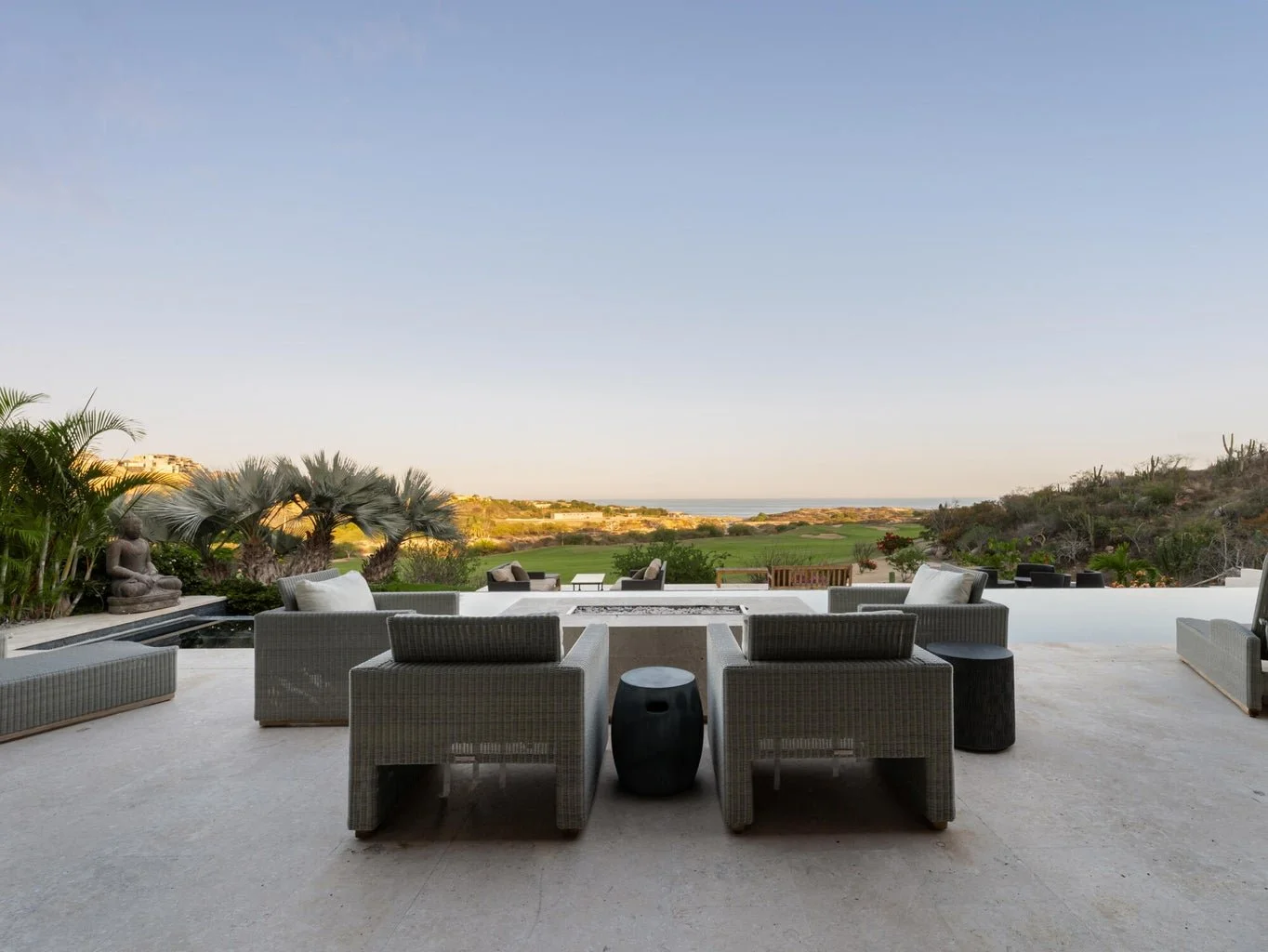 Outdoor patio with wicker furniture, cushions, and small tables overlooking a green landscape, hills, and ocean under a clear sky.