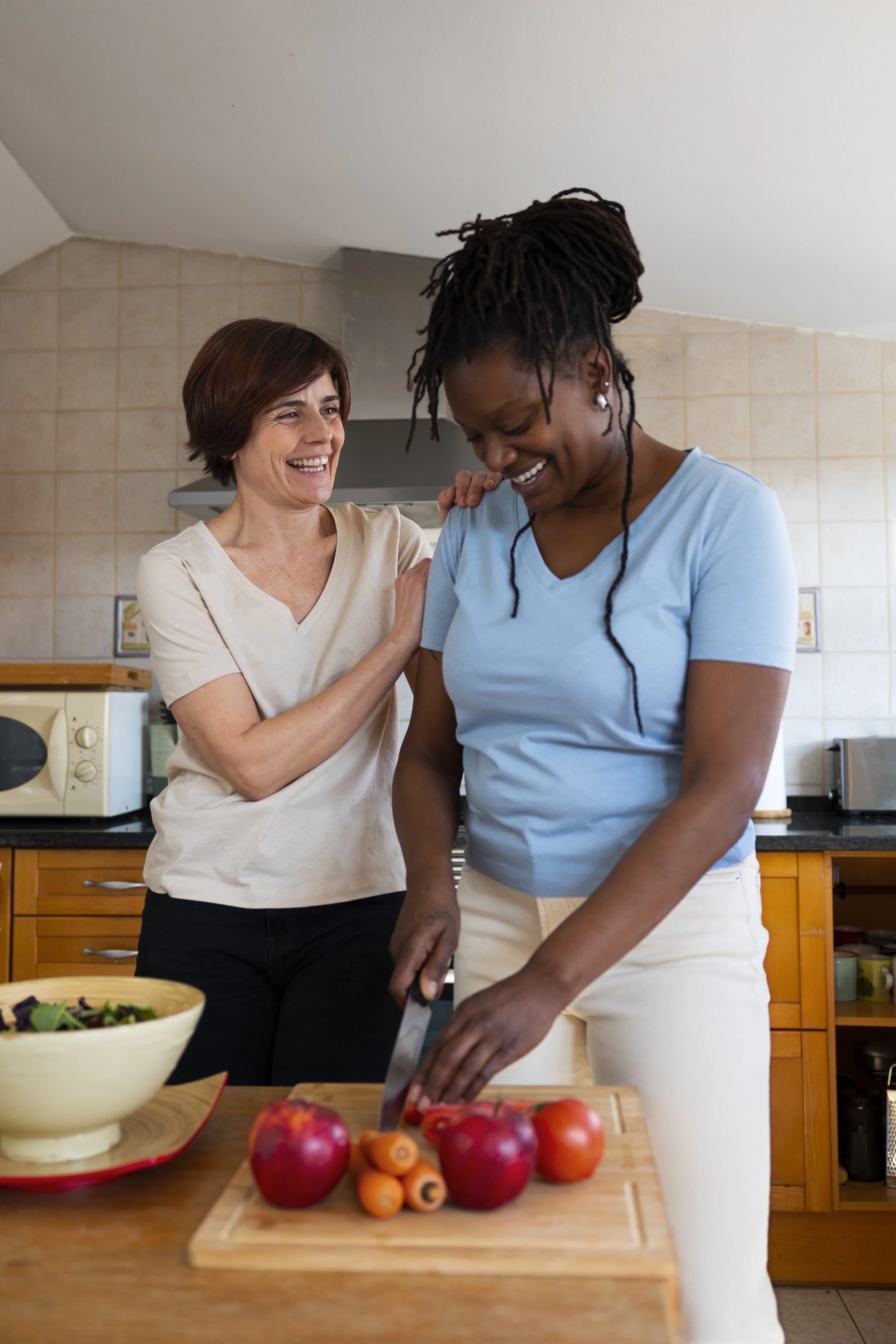 Two women smiling and preparing vegetables in a kitchen. One woman is chopping carrots and tomatoes on a cutting board, while the other woman watches and smiles.