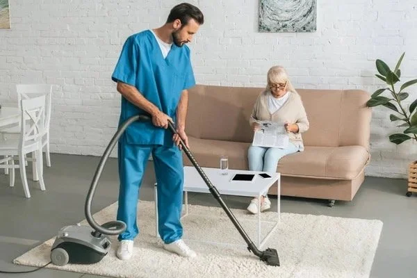 A man in blue scrubs vacuuming a living room with an elderly woman sitting on a beige sofa reading a newspaper.