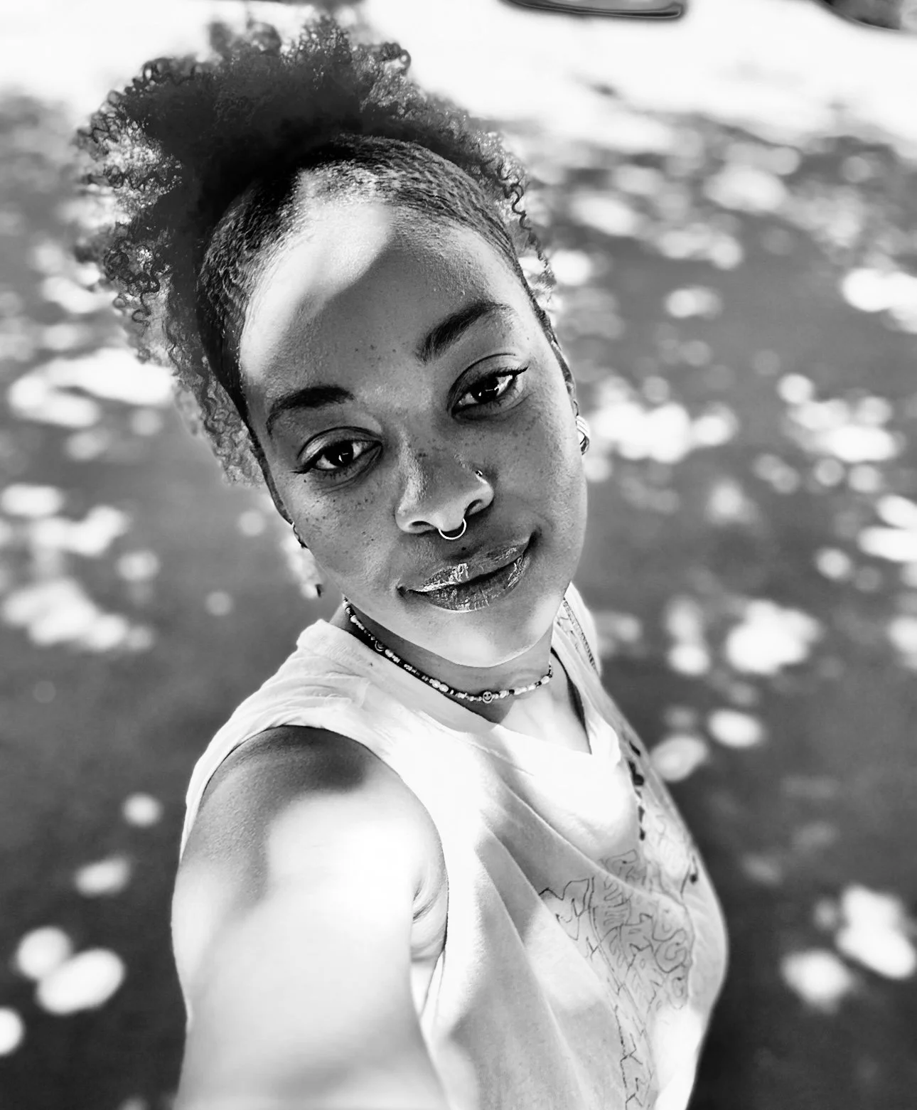A young woman with curly hair styled in a high puff taking a selfie outdoors in black and white. She has facial piercings, wears jewelry, and is smiling slightly while looking at the camera. The background appears to be a body of water with light reflections.