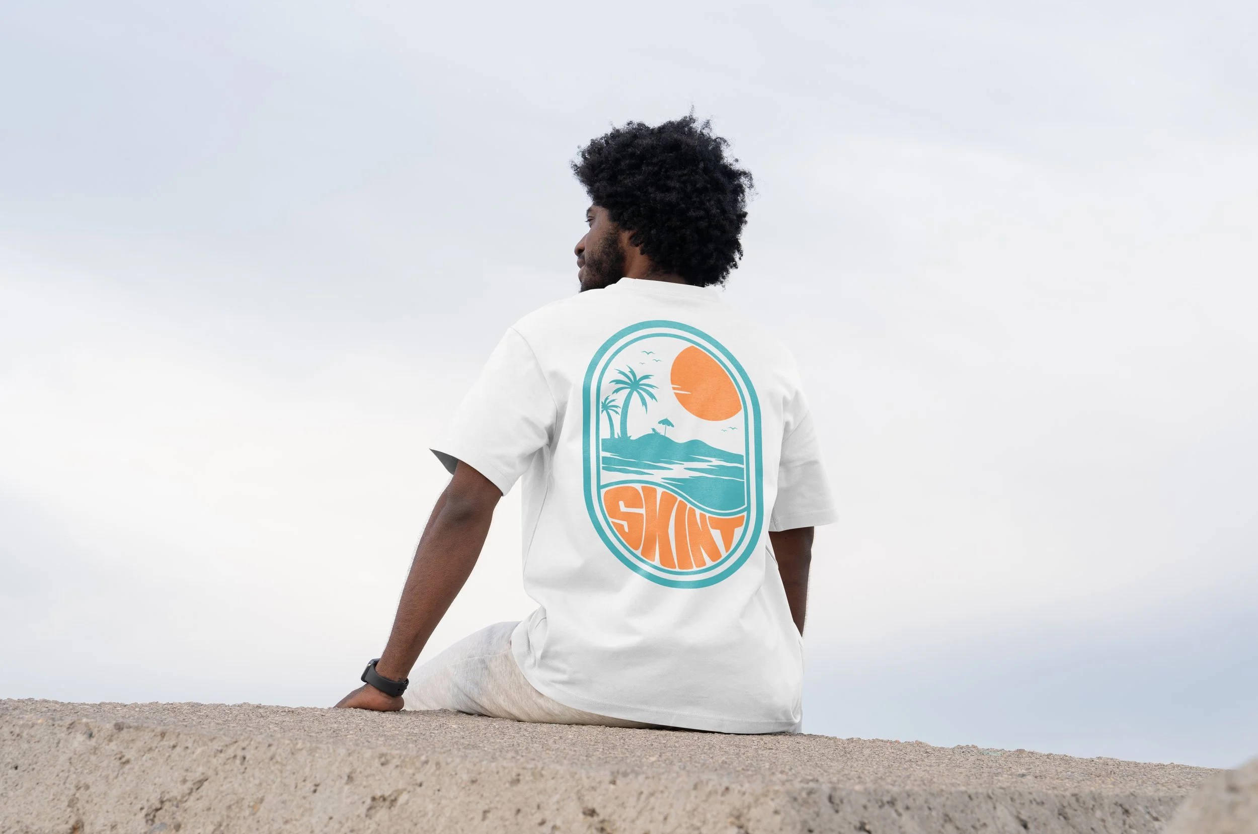 Man sitting on sand beach wearing a white T-shirt with a graphic of a tropical scene and the word "SKINT" on the back