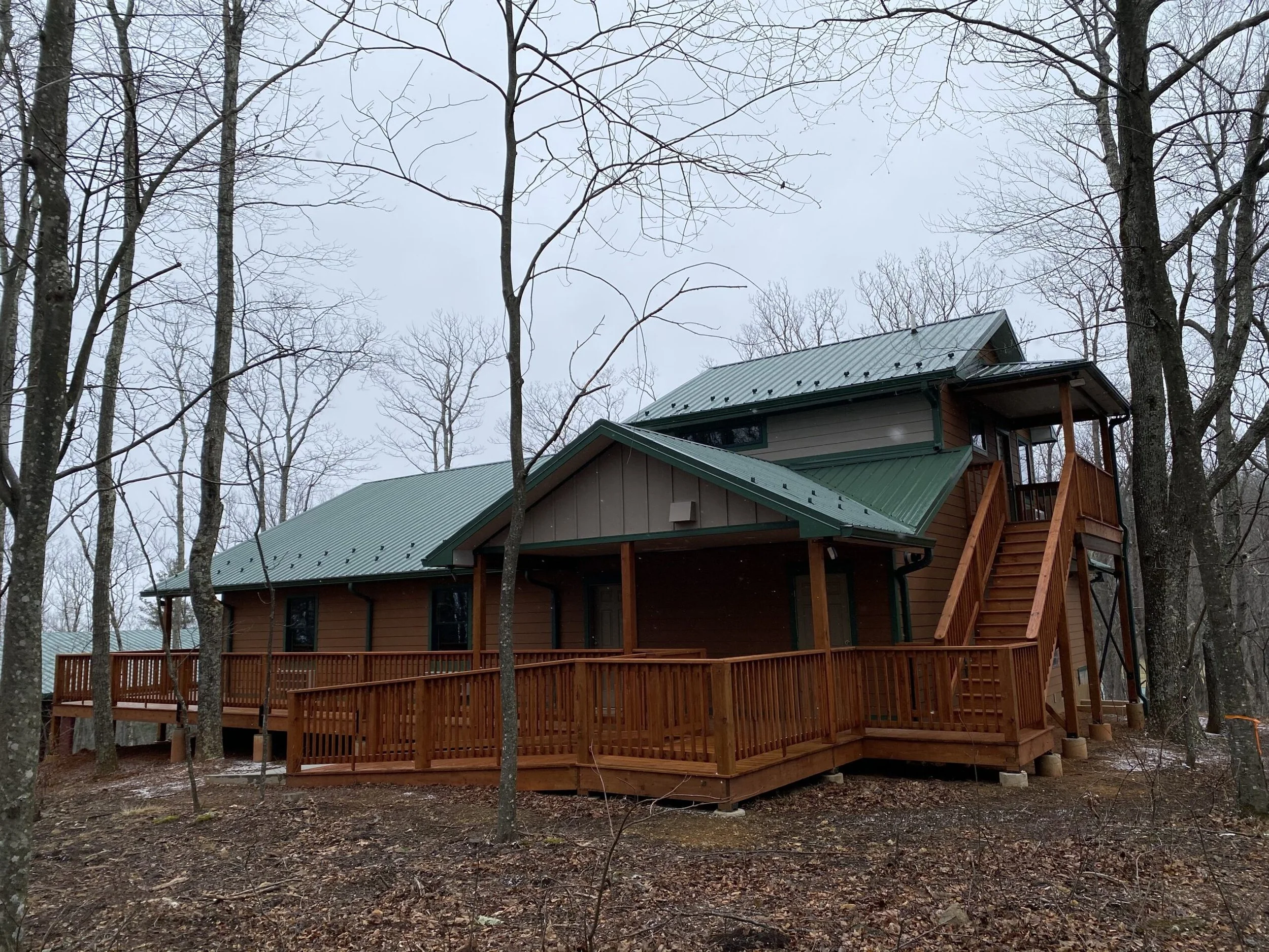 A multi-story house with a green metal roof and wooden exterior, surrounded by leafless trees, with a large wooden deck and staircase, in a wooded area during winter.