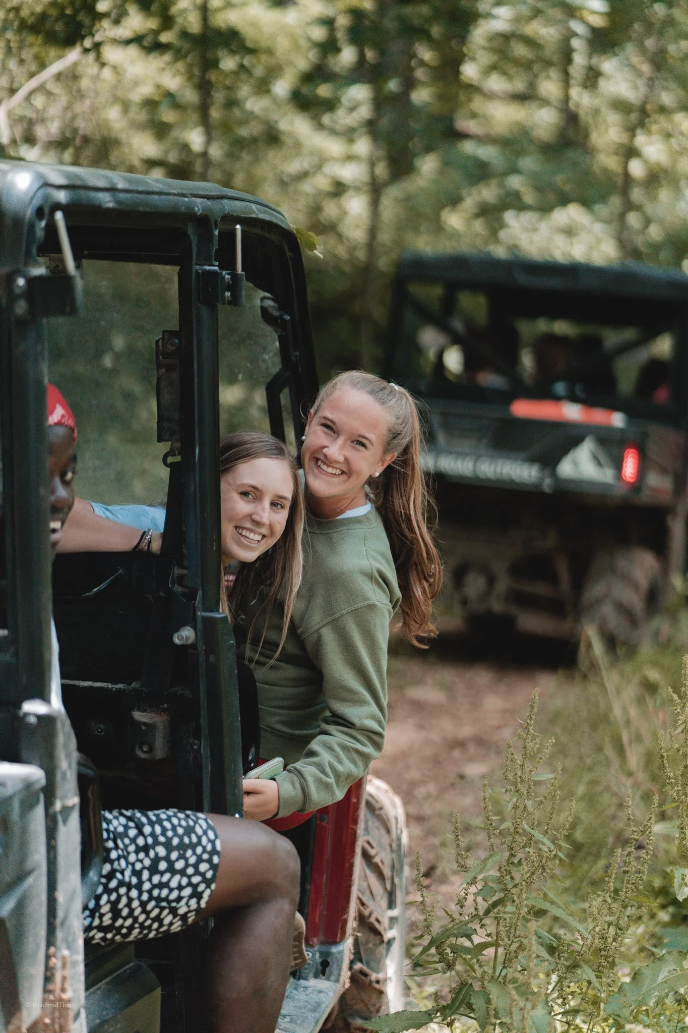 Two smiling girls leaning out of an off-road vehicle in a wooded area, with another vehicle in the background.