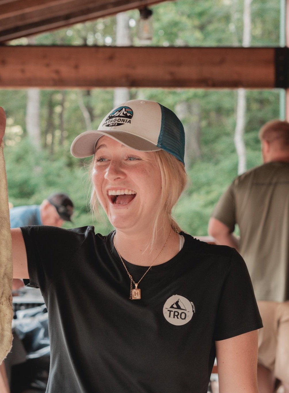 A young woman wearing a Patagonia cap and a black T-shirt with a mountain logo is smiling and laughing with an outdoor, wooded background.