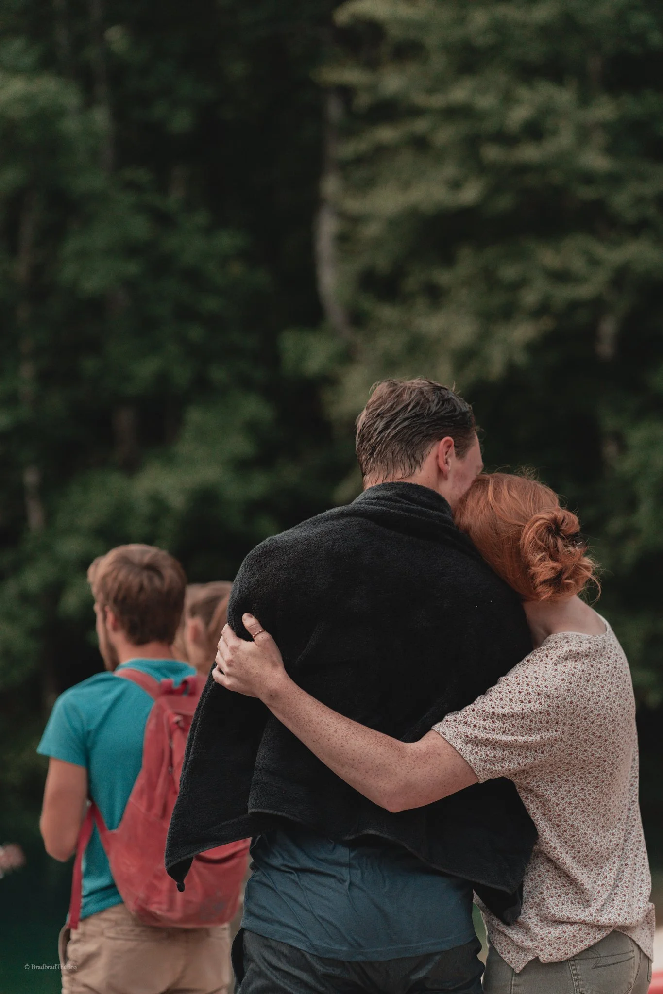 A couple embracing outdoors in front of a forest, with two young men and a woman in the background.