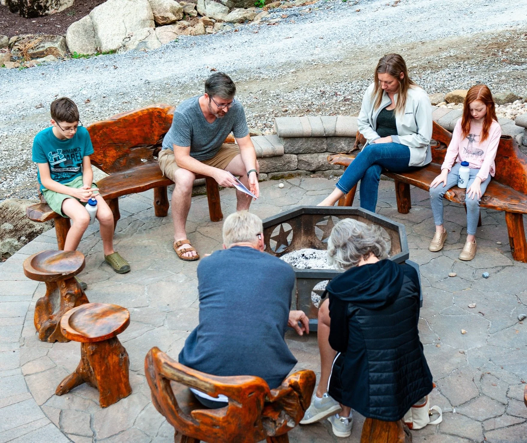 A group of six people, including children and adults, sitting around a fire pit with wooden benches, outdoors on a paved stone area, with gravel and rocks in the background.
