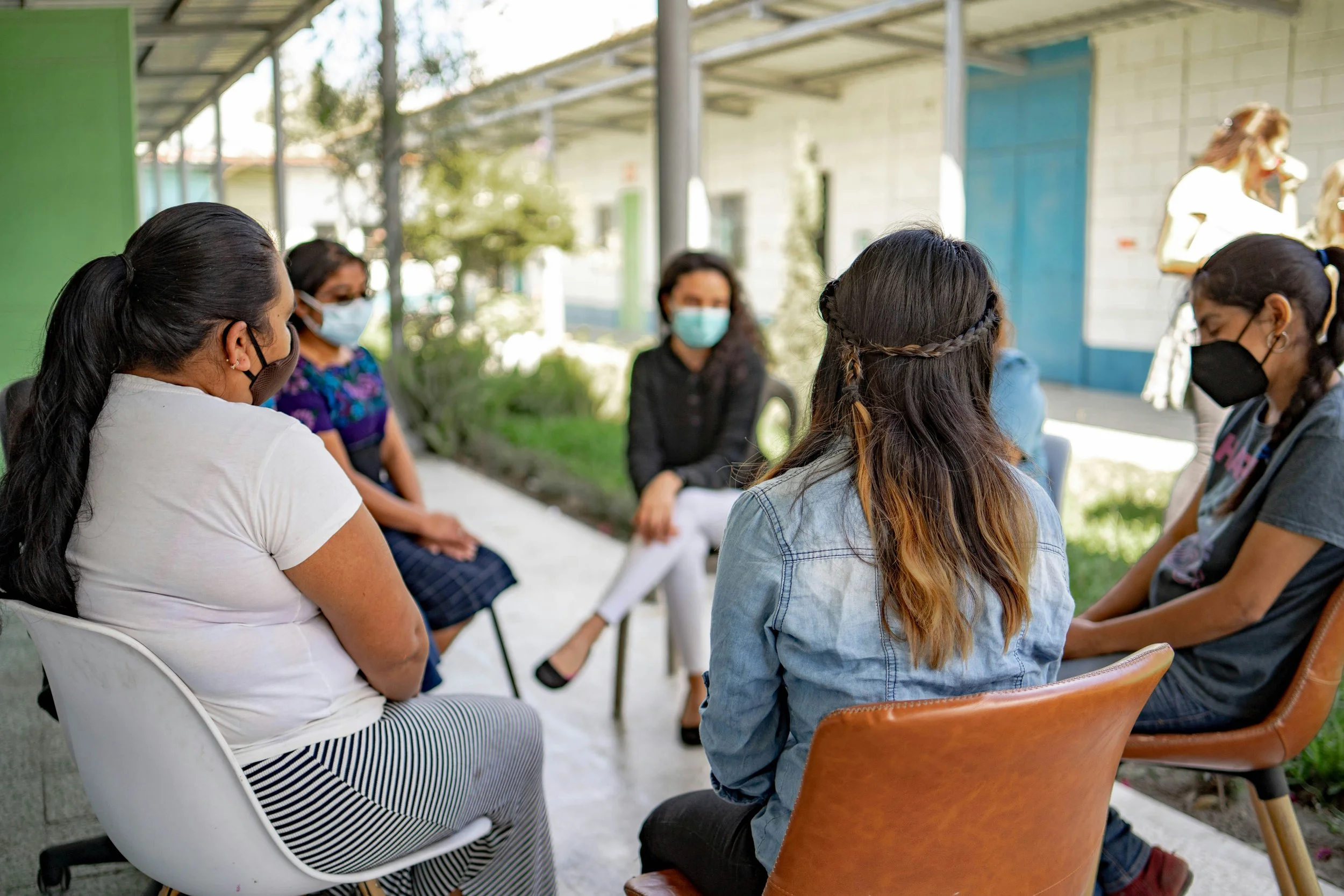 Masked women gathered in an outdoor community meeting, seated in a circle and talking together.