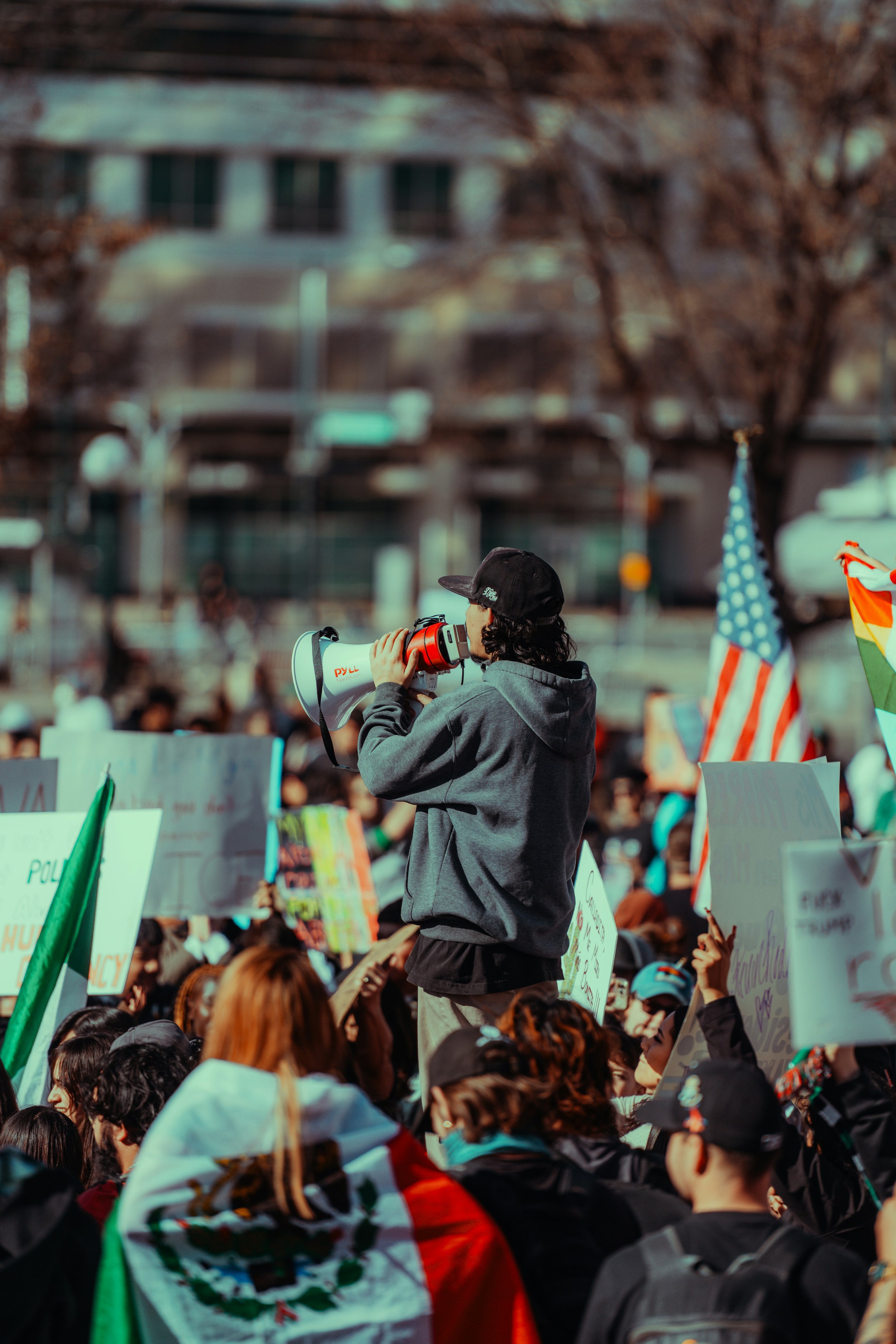 Protester in a hoodie and cap speaks into a megaphone above a large outdoor crowd holding signs and Mexican and American flags.