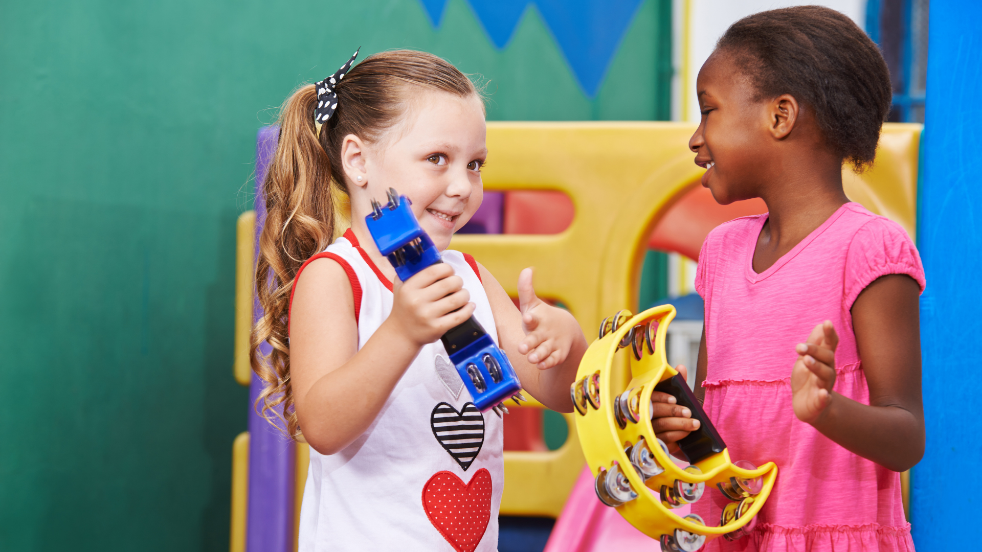 Two young girls playing with musical instruments in a colorful indoor play area, smiling and engaging with each other.