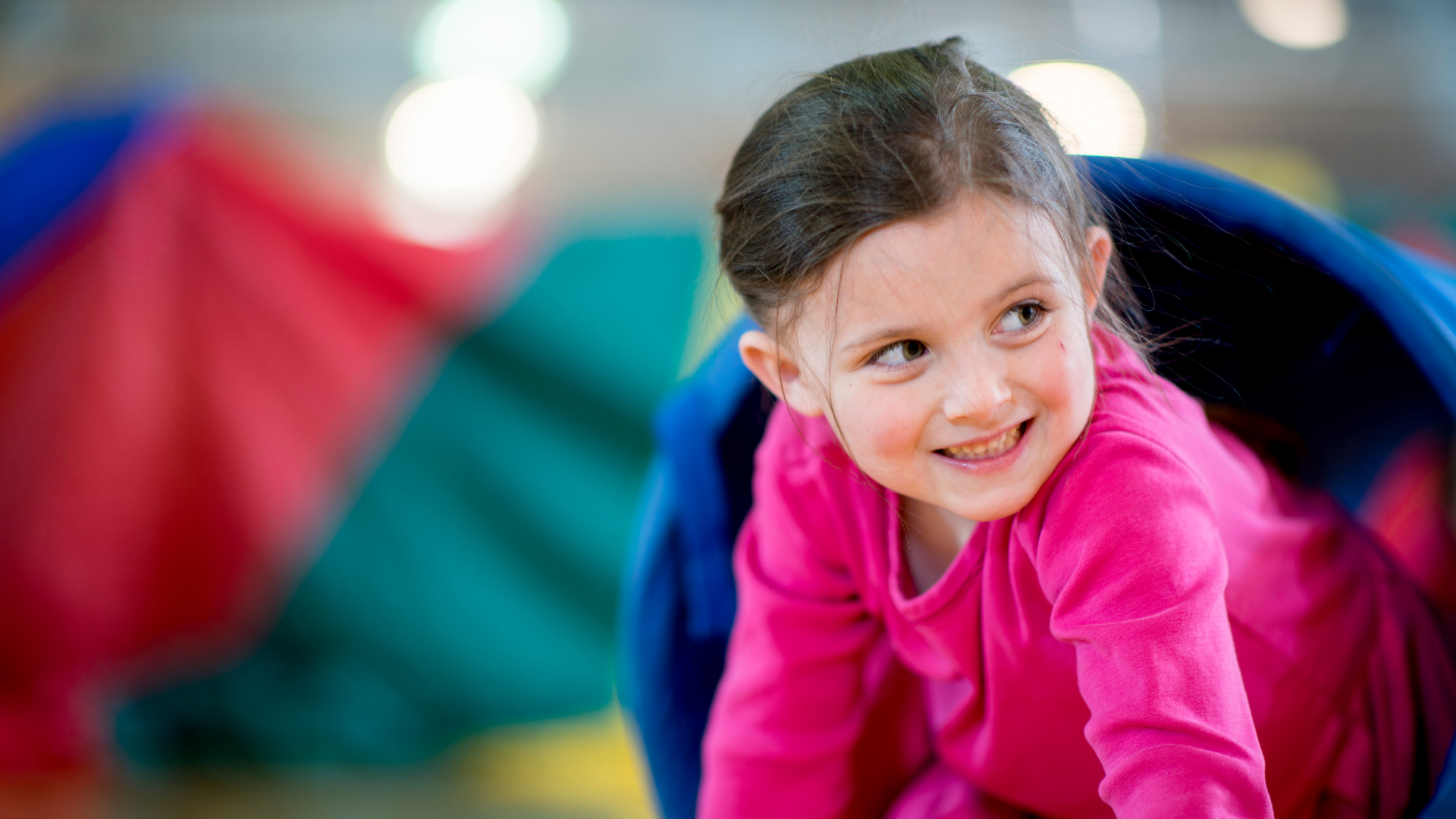 Young girl in a pink shirt crawling out of a colorful play tunnel, smiling and looking to the side.