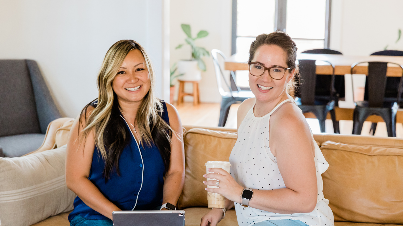 Two women smiling on a beige couch in a bright living room or cafe, one holding iced coffee, with glass doors and dining table in background.
