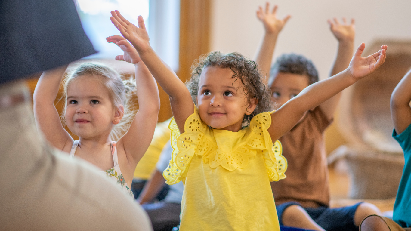 Group of young children sitting on the floor and raising their hands during a classroom activity.