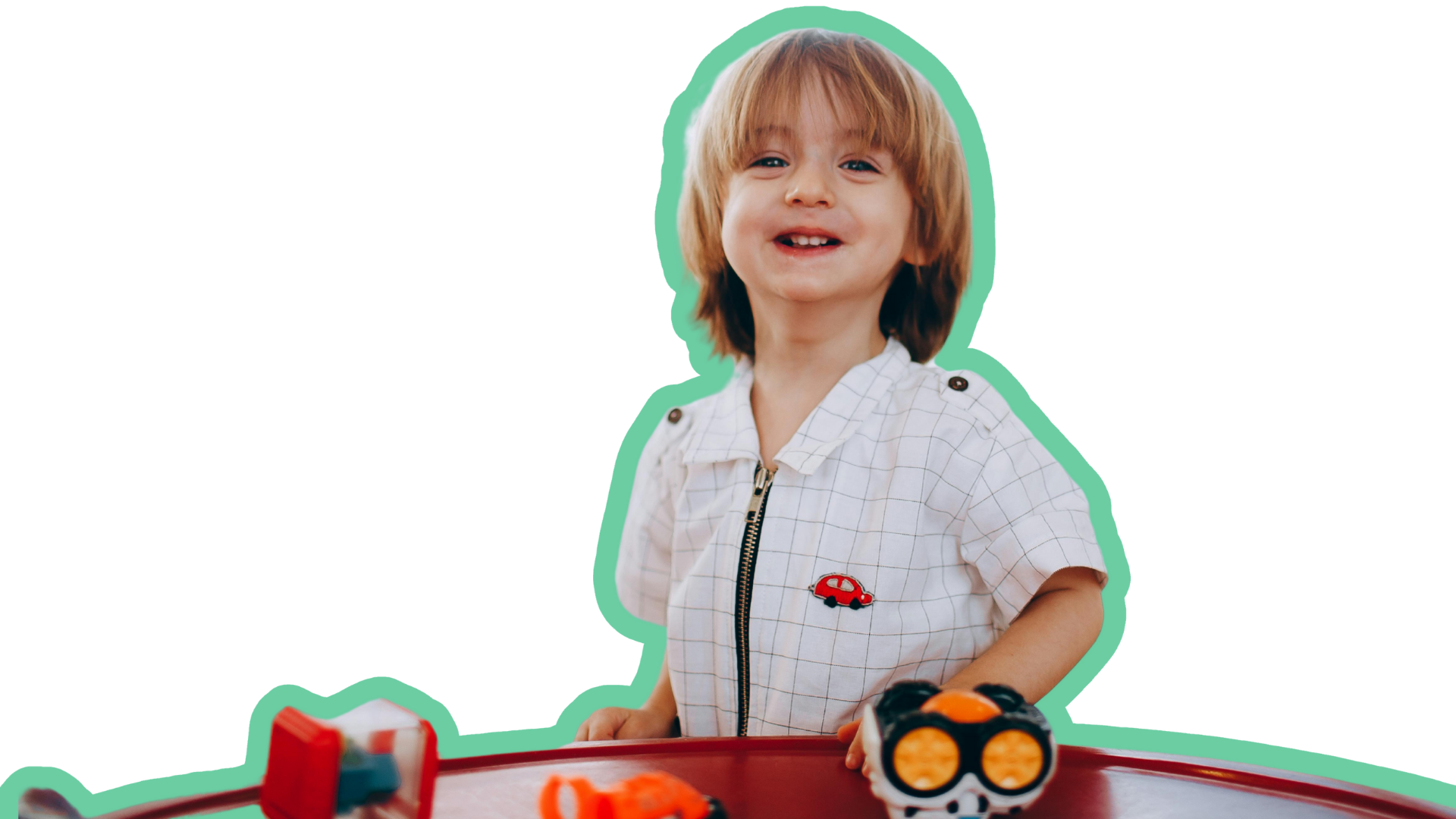 A young boy with blond hair, smiling and wearing a white shirt with a small red car patch, playing with toy cars on a red table.