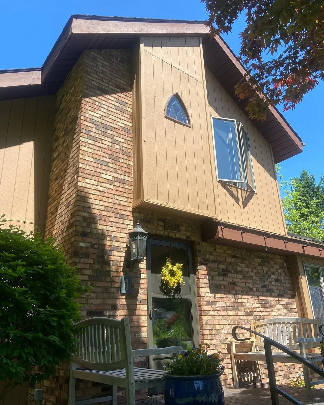 A two-story house with brick lower level and wood-paneled upper level, featuring a brick chimney, a hanging yellow wreath door decoration, an outdoor lantern, and a patio with a wooden bench and flower pot. Visible trees and blue sky in the backgroun
