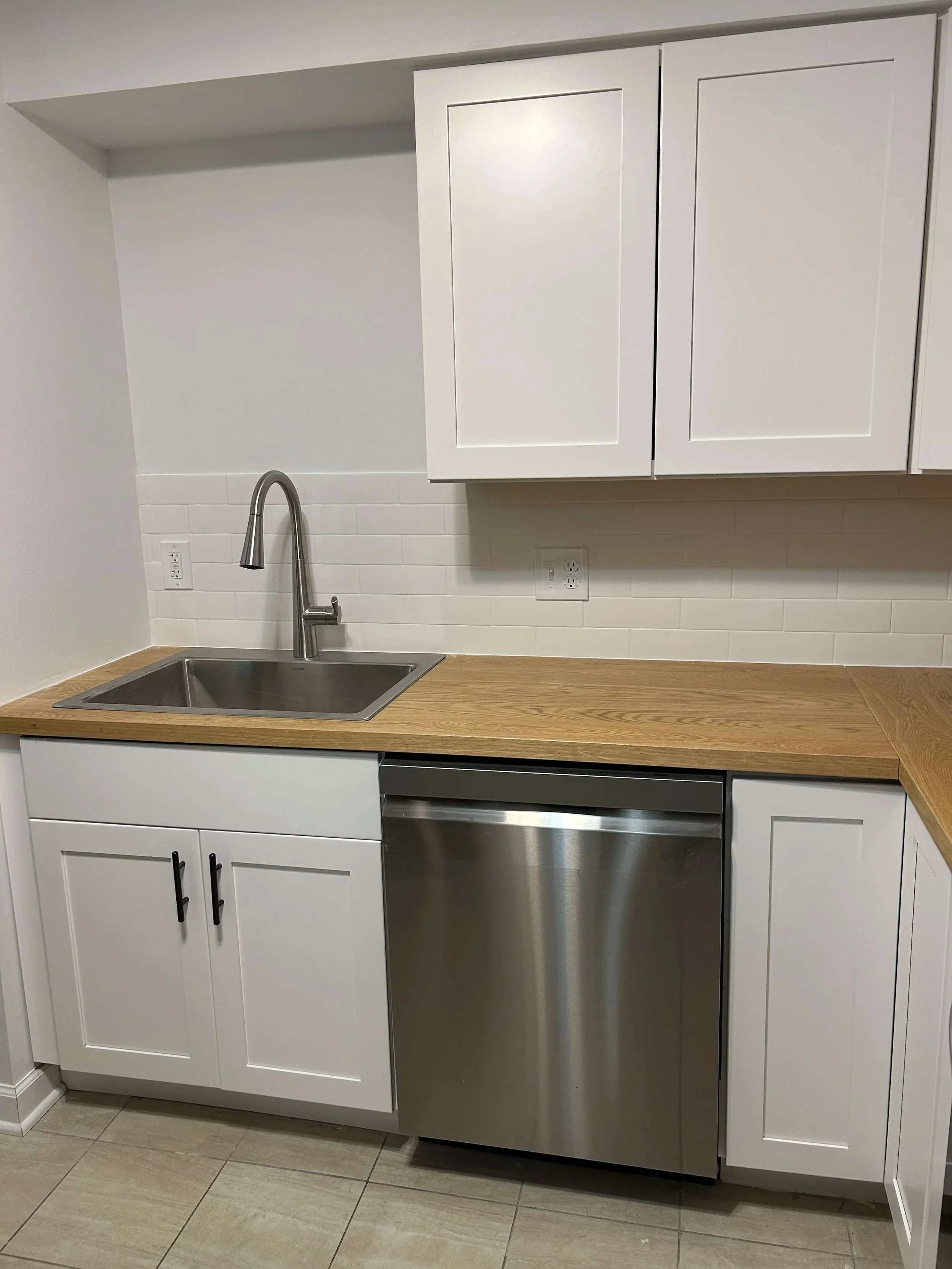 Kitchen countertop with stainless steel Kohler sink, white cabinets, and oak veneer countertop, white backsplash, and electrical outlets.
