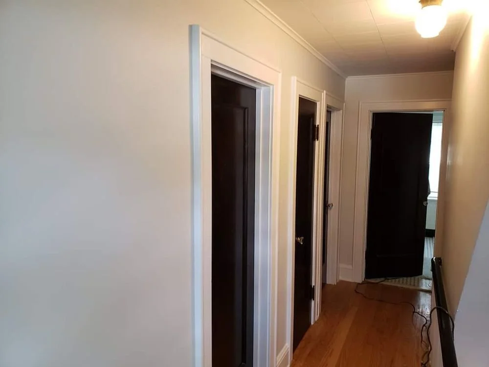 A hallway with three closed dark wooden doors and a fourth door slightly open at the end, with a light-colored wall on the left, a wooden floor, and ceiling lighting.
