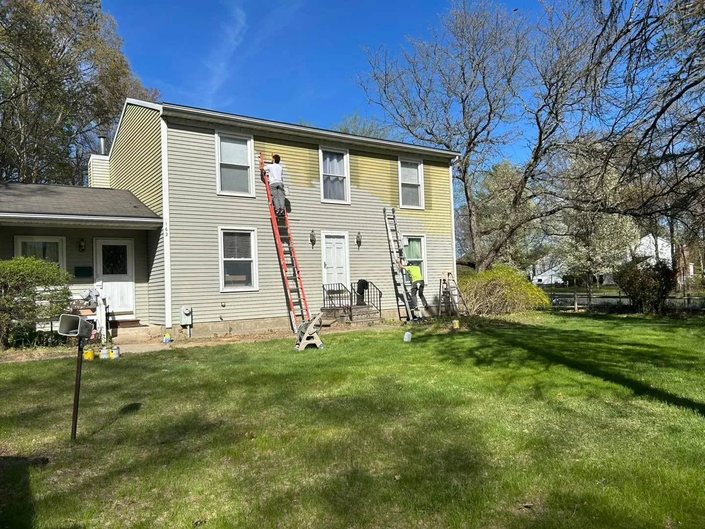 People working on the exterior of a two-story yellow house with white trim in a backyard with green grass and trees.