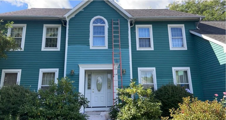 A two-story blue house with white trim, multiple windows, a front door, and a ladder leaning against the upper part of the house.