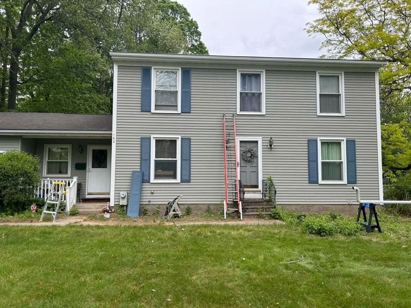 Exterior painting project on two-story gray house with white trim, blue shutters, and ladder on green lawn.