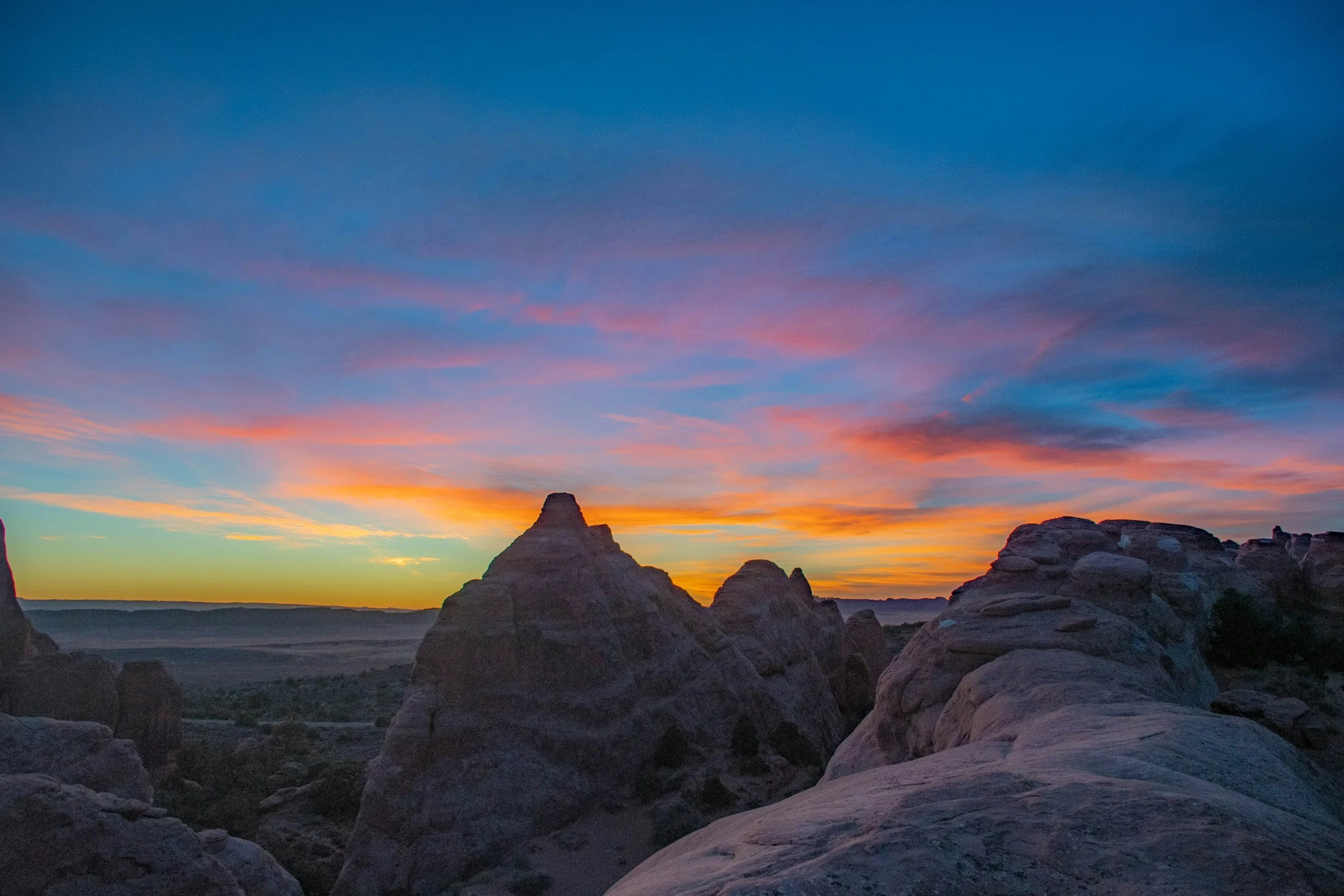 Sunset over rocky desert landscape with layered rock formations and colorful sky with pink, orange, and blue clouds.