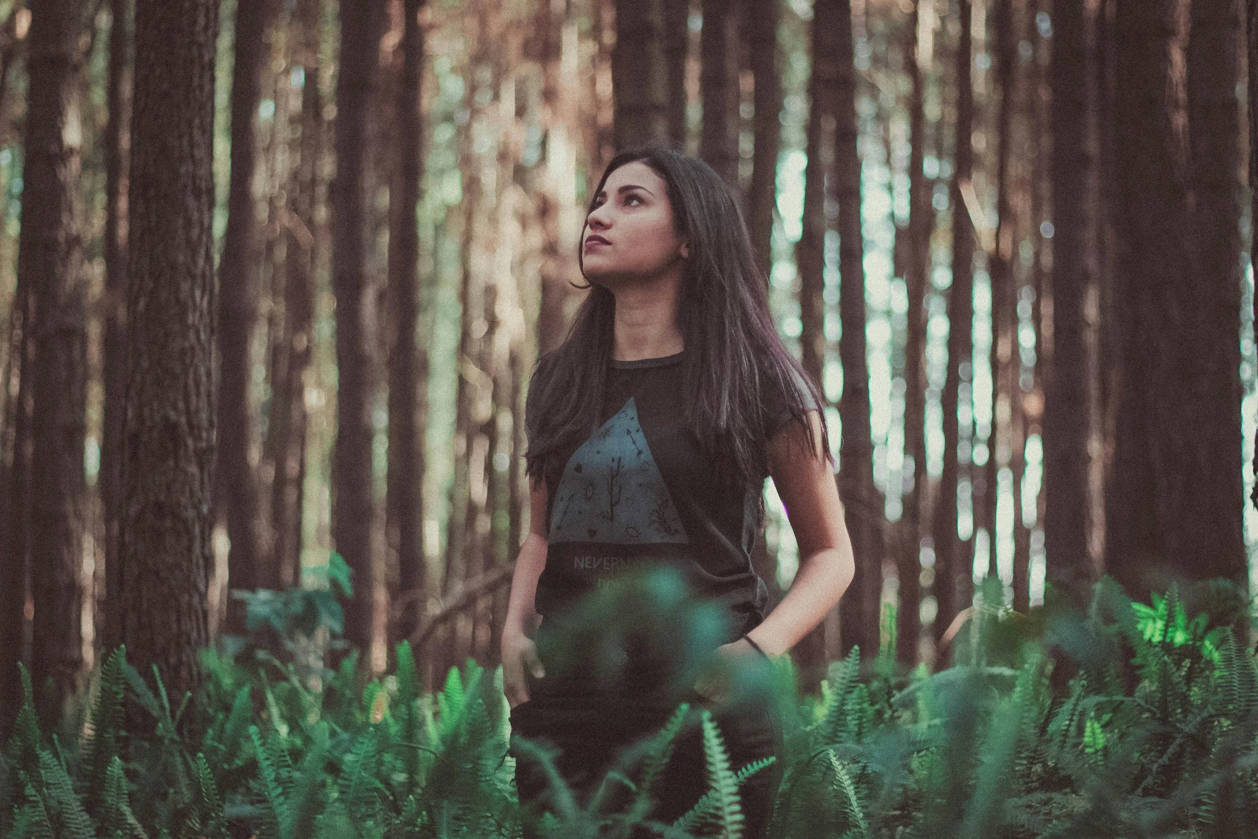 A young woman with dark hair standing in a forest among tall trees and green ferns, looking up with a thoughtful expression.