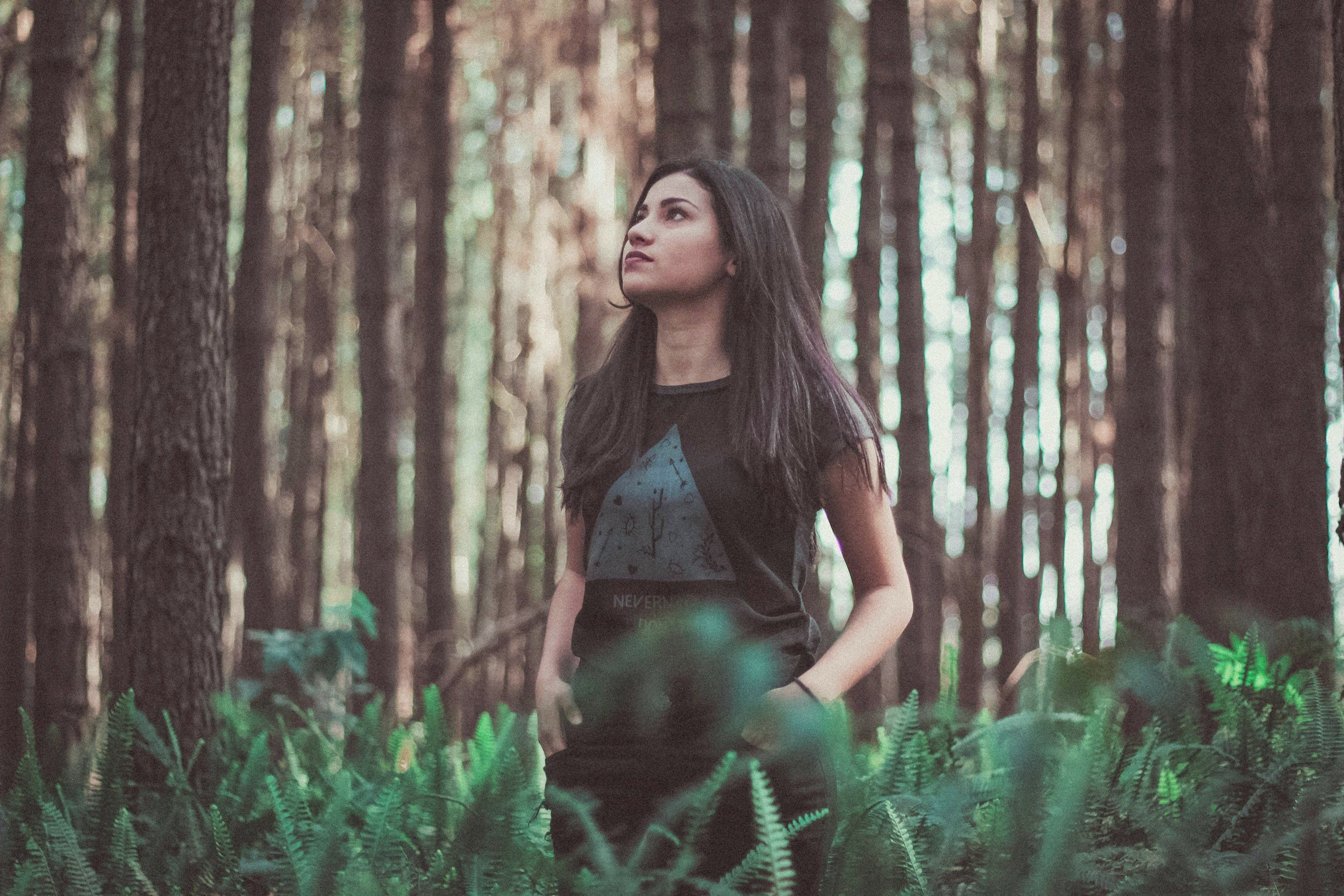 Young woman with long dark hair standing in a dense forest of tall trees with green foliage, looking upwards