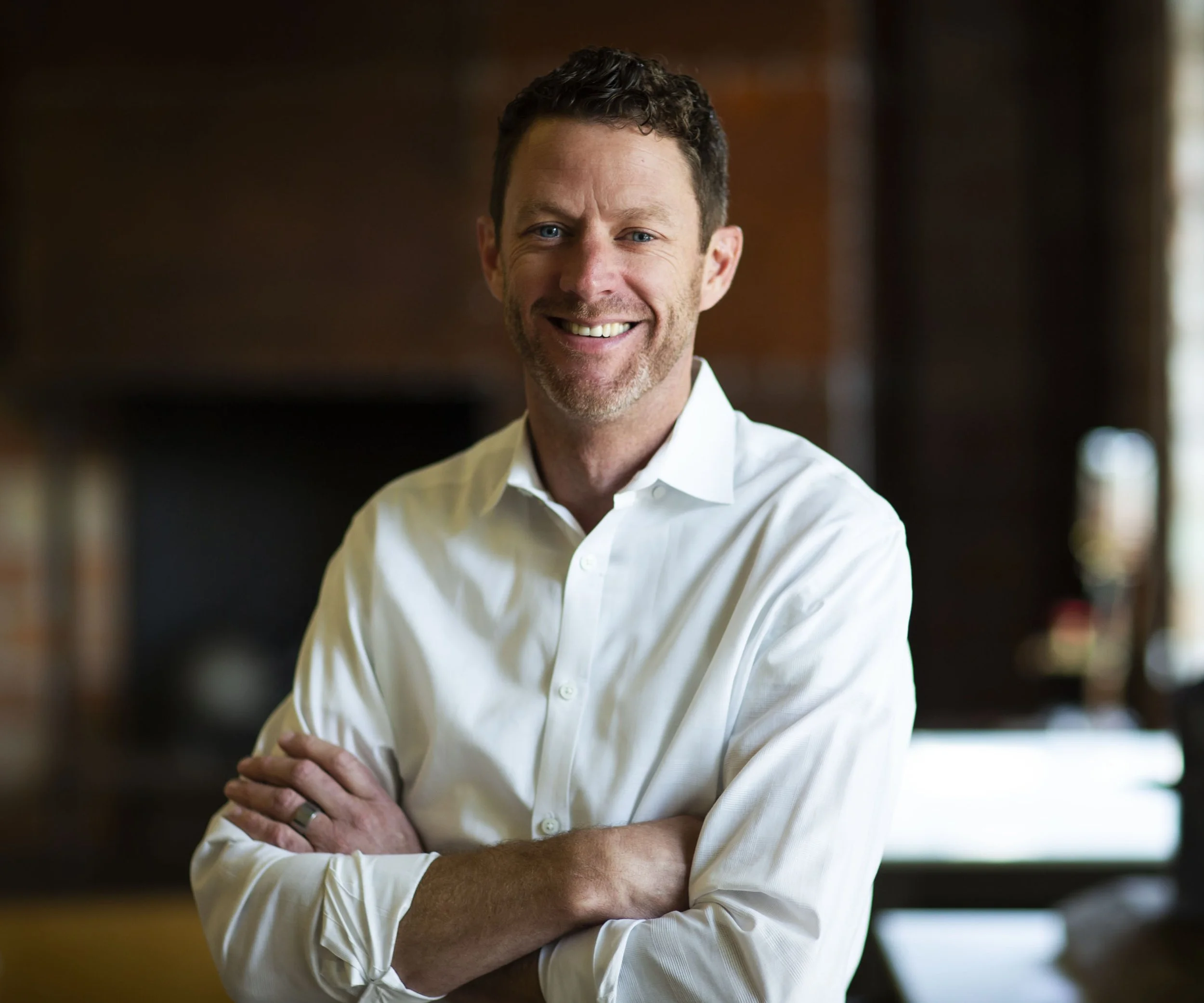 A smiling man with short dark hair and blue eyes, wearing a white button-down shirt, standing with arms crossed in a warmly lit room.