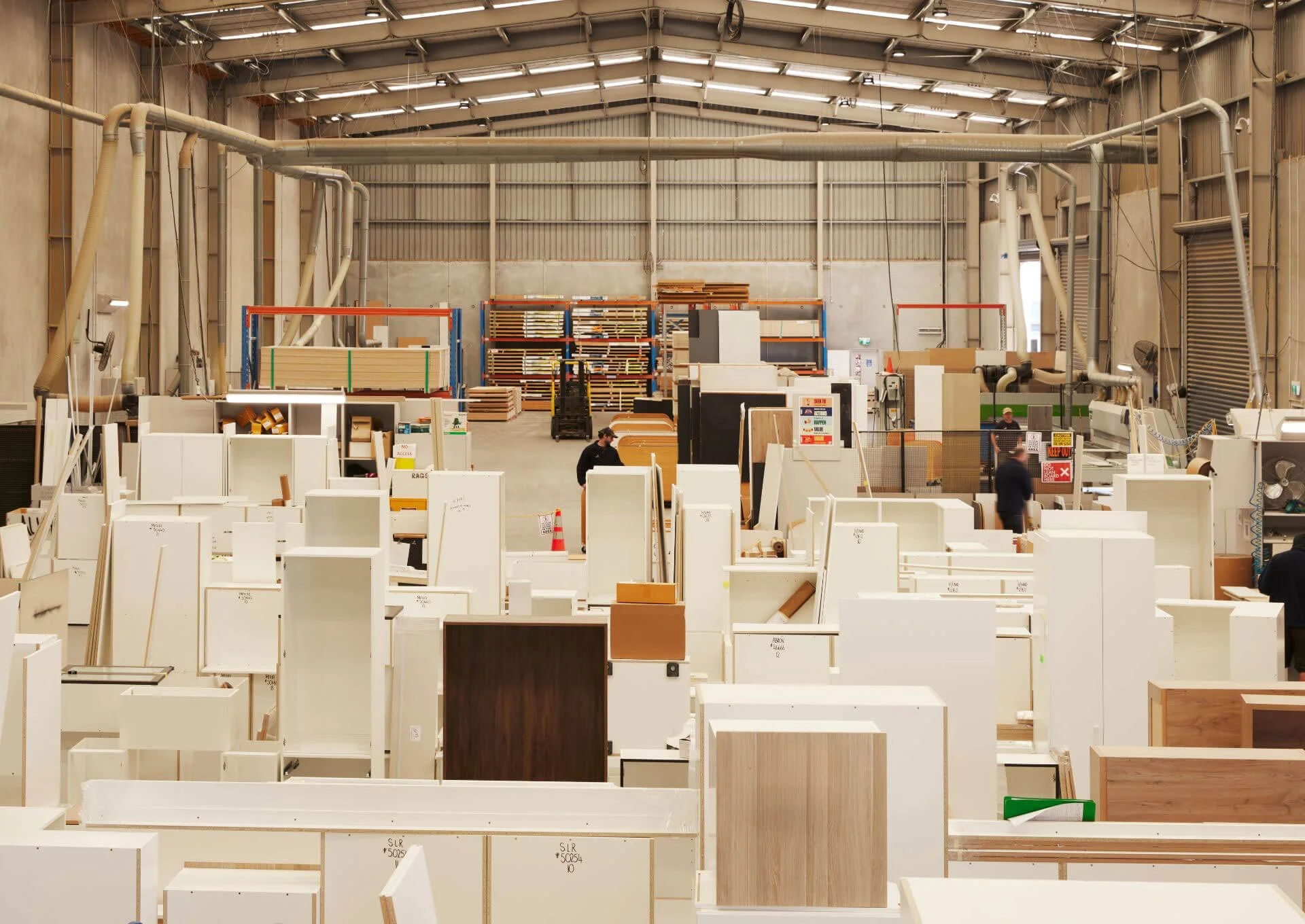 A large warehouse with industrial shelving filled with wood planks and furniture components, and workers assembling furniture.
