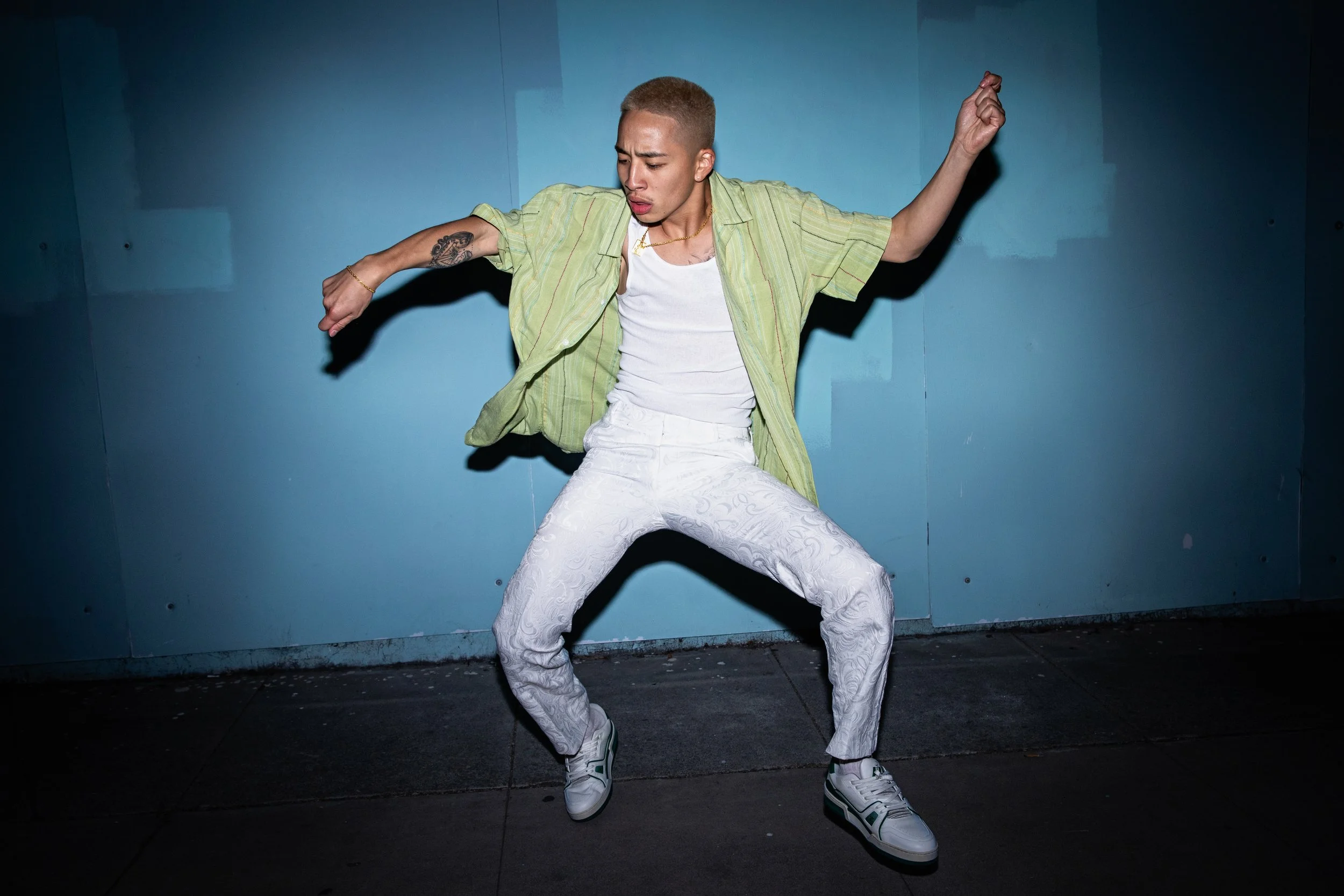 A man dancing against a blue wall, wearing a green short sleeve button-up shirt, white patterned pants, and white sneakers.