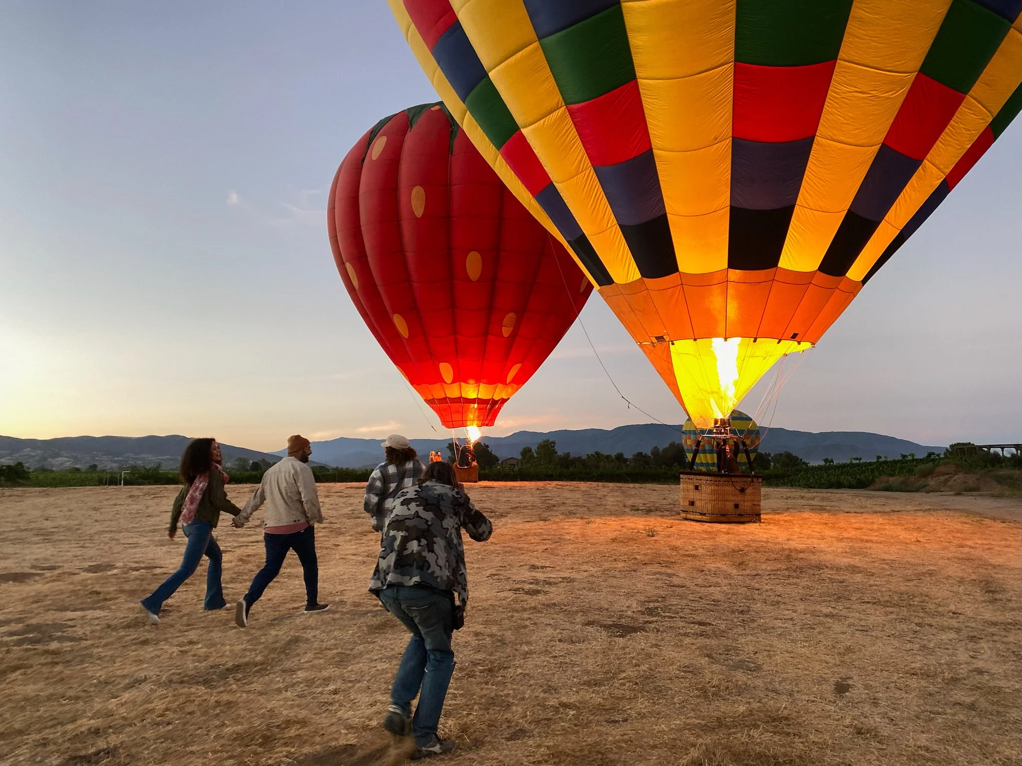 Four people are running towards two large colorful hot air balloons preparing for takeoff on a dry field during sunset.