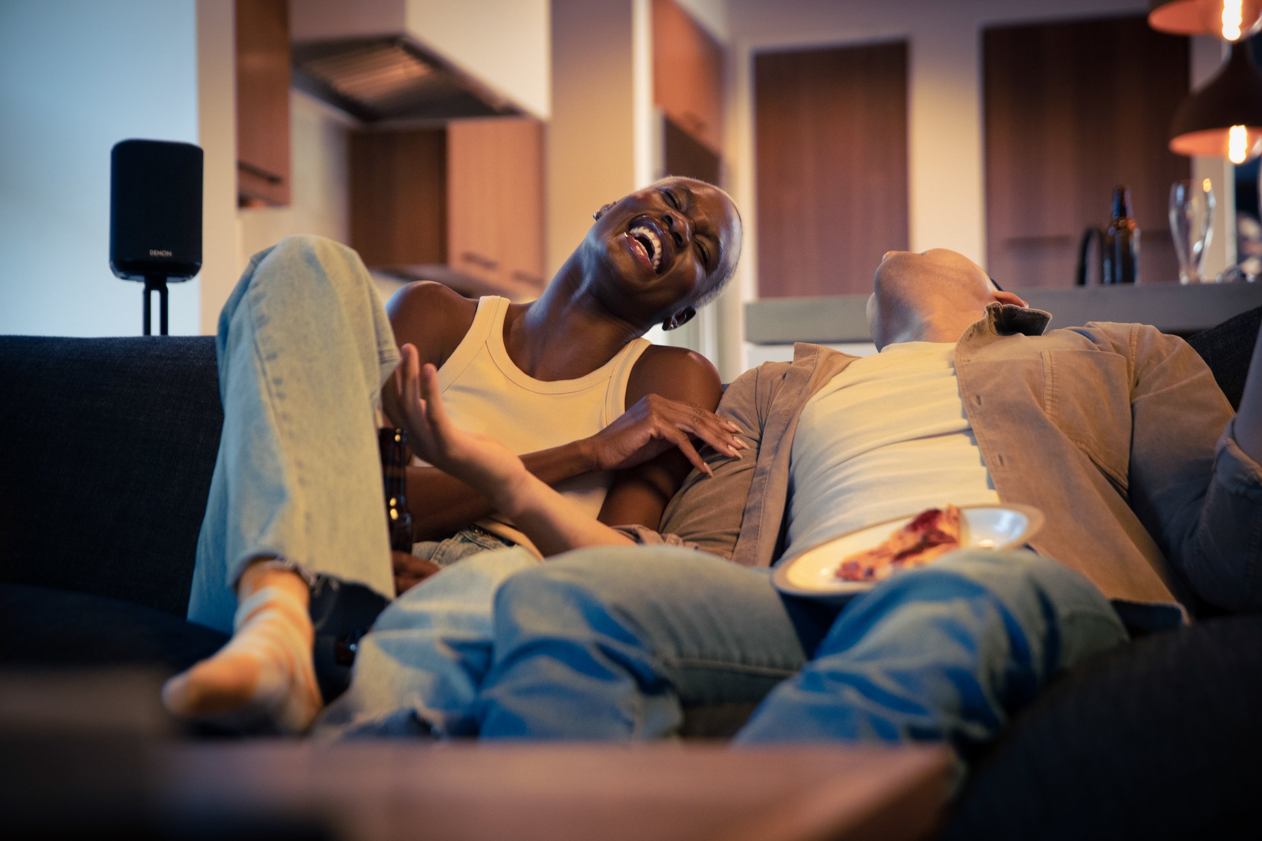 Two people are sitting on a couch, laughing and having a good time, with a pizza on a plate in front of them in a cozy living room.