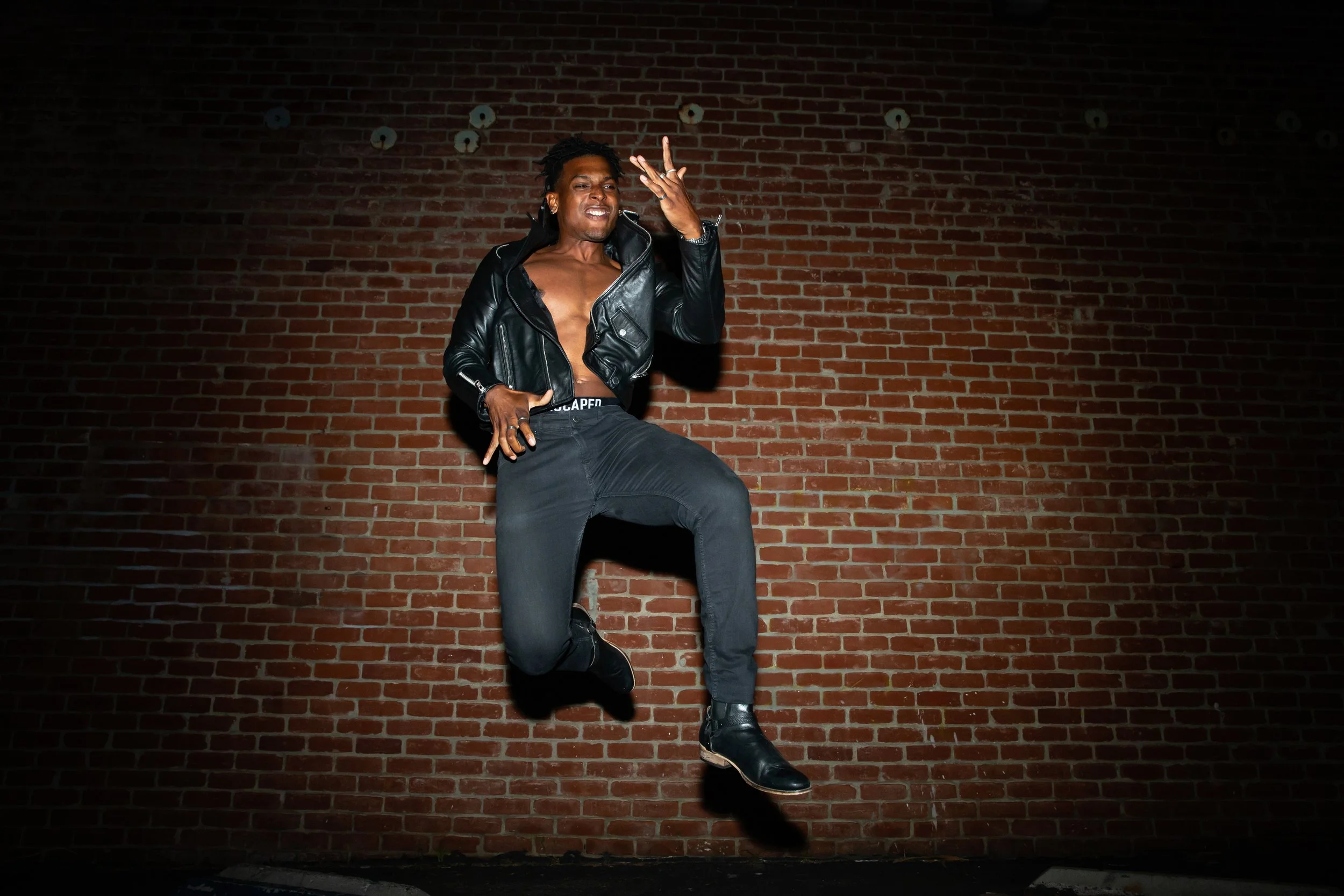 A man dance jumping in front of a red brick wall wearing a black leather jacket, black pants, and black boots.
