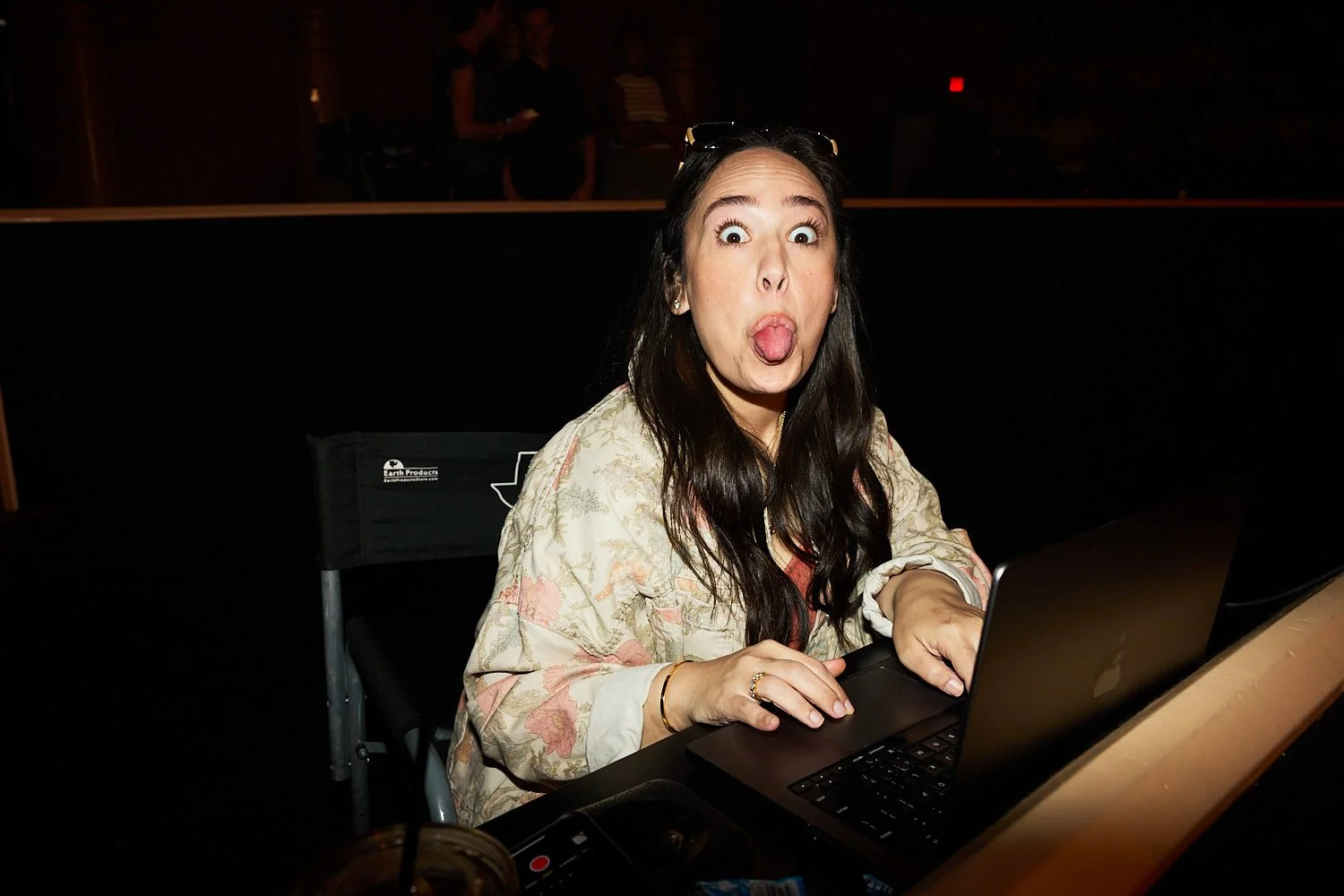 A woman with long dark hair making a funny face, sticking out her tongue, while sitting at a table with a laptop.