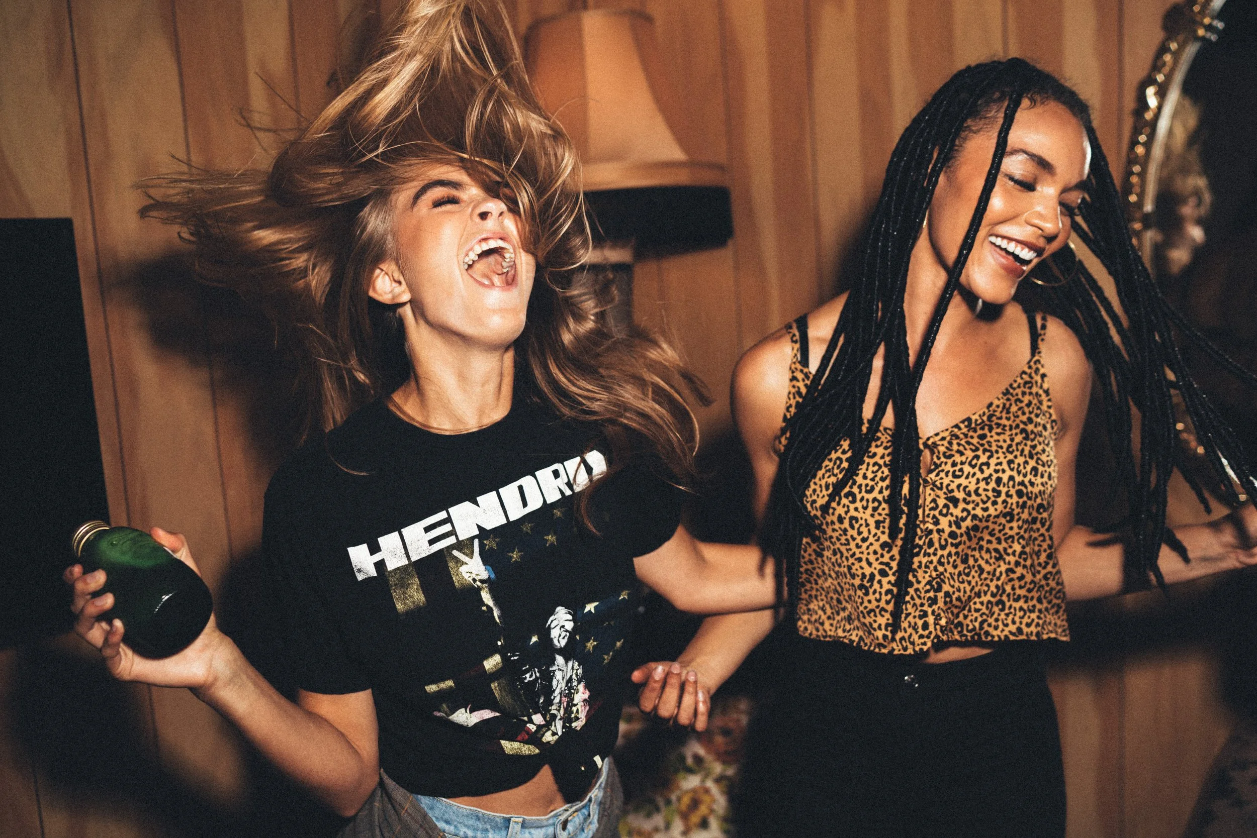 Two women are dancing and laughing joyfully at a party or social gathering. One woman has blonde hair, wearing a black graphic t-shirt, holding a green bottle, and her hair is flying around her face. The other woman has long black braided hair, wearing a leopard print sleeveless top. They are inside a room with wooden panel walls, creating a lively and fun atmosphere.