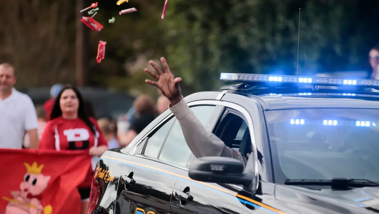 A person in a police car waving out the window, with a parade and people holding a pink pig-themed banner in the background.