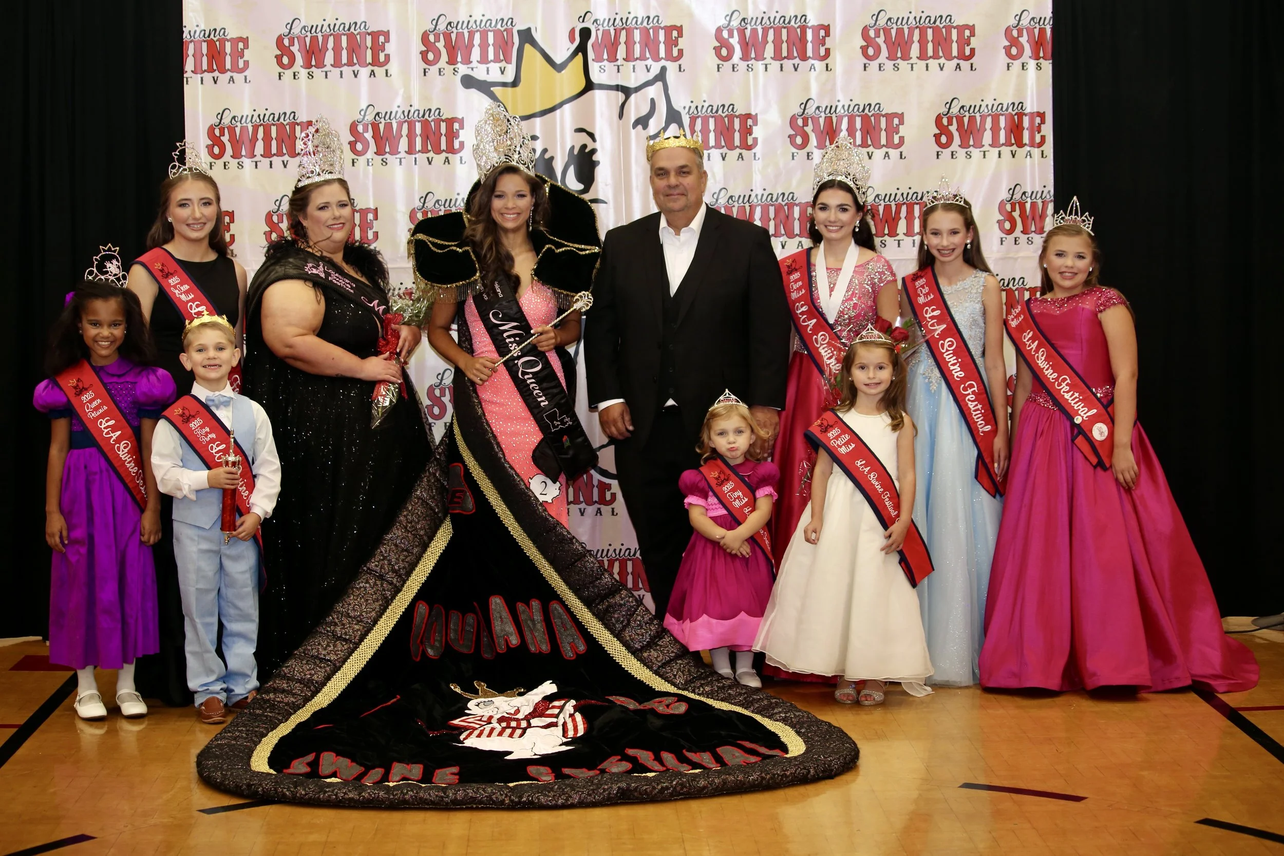 Group of young girls and women in fancy dresses and sashes celebrating at a festival, with some wearing crowns, standing in a barn-like structure, with a large black and red quilt or banner in front that says 'Sunshine Festival' and features a cat mascot.