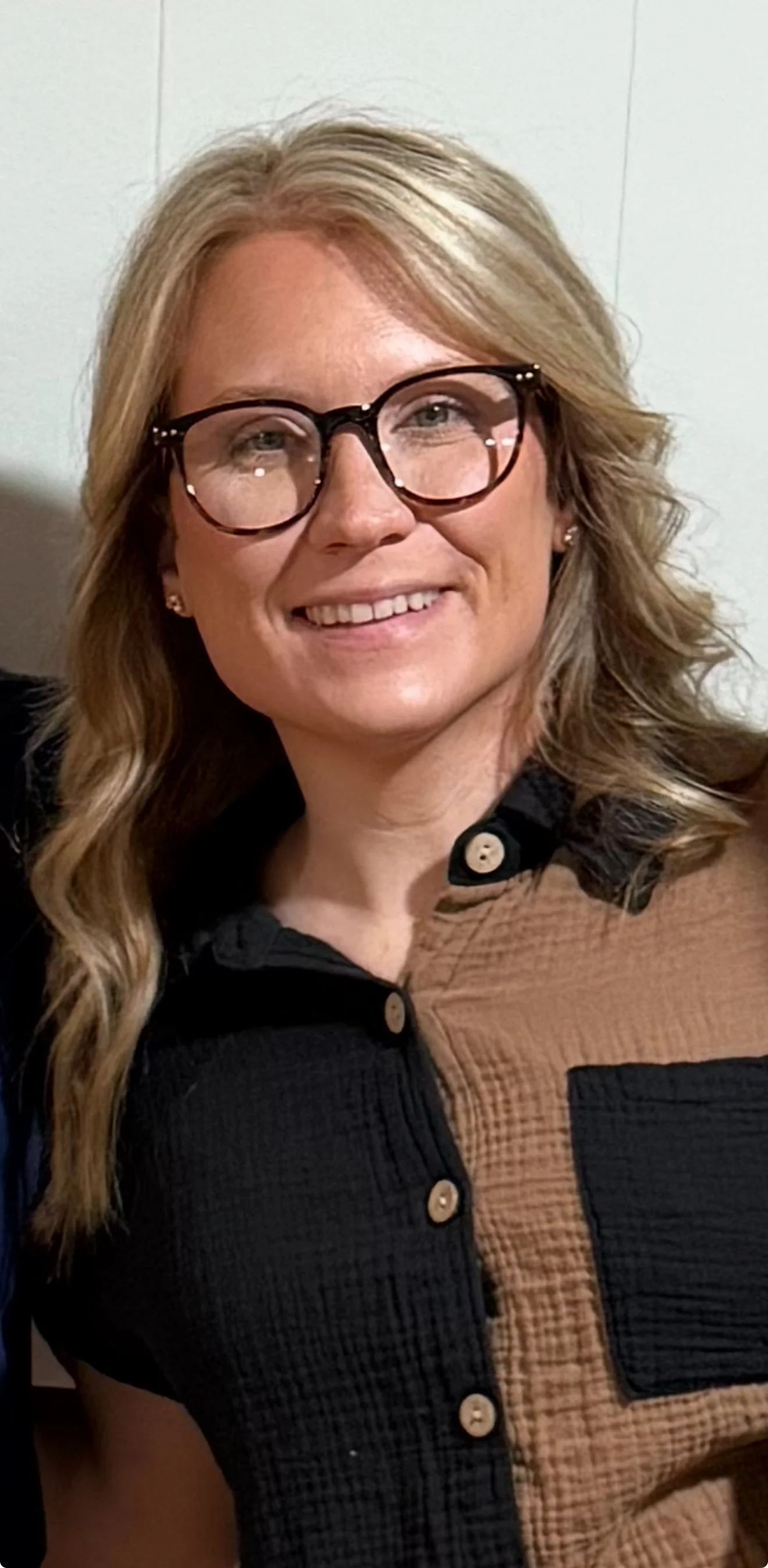 A woman with blonde wavy hair, wearing glasses, smiling, dressed in a color-blocked shirt with black and brown sections and beige buttons.