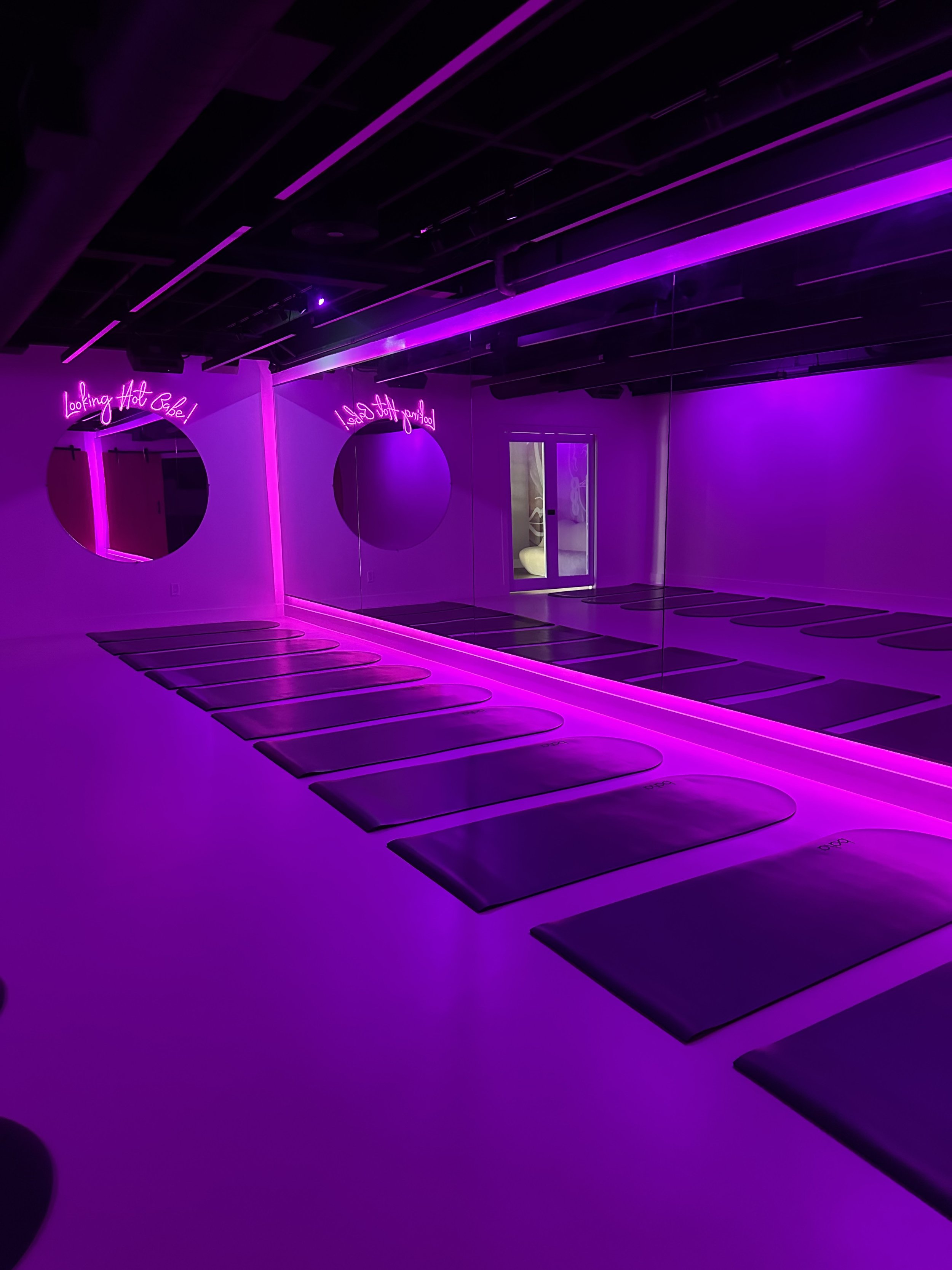 Yoga studio with black mats illuminated by purple neon lights, mirrors on the wall, and neon signs that say 'Looking Hot Babe!' and 'I Feel Confident'.