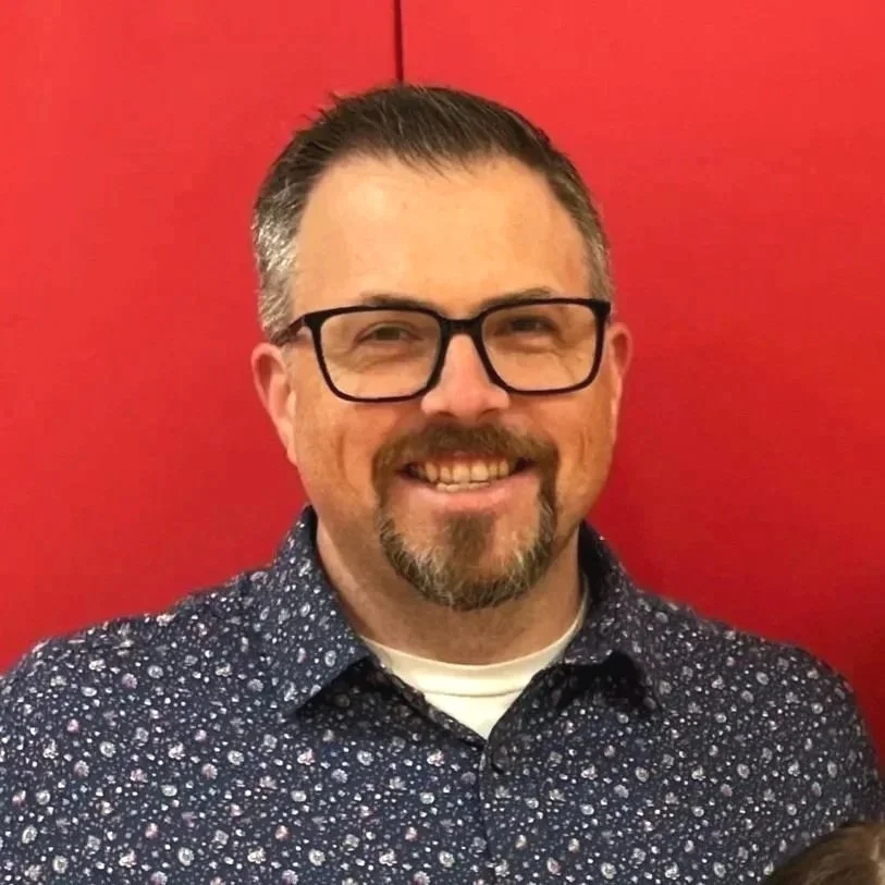 Man in glasses with beard and mustached, smiling with a patterned button up shirt in front of a red wall.