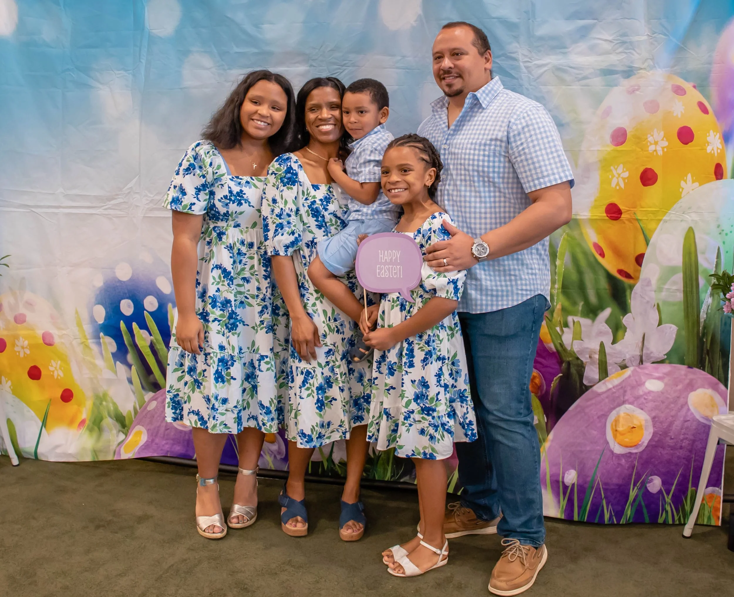 Family celebrating Easter in front of a colorful backdrop with decorated eggs and spring flowers. The women and girls are wearing matching blue and white floral dresses, while the man and boy are dressed in casual shirts and jeans. One girl holds a s