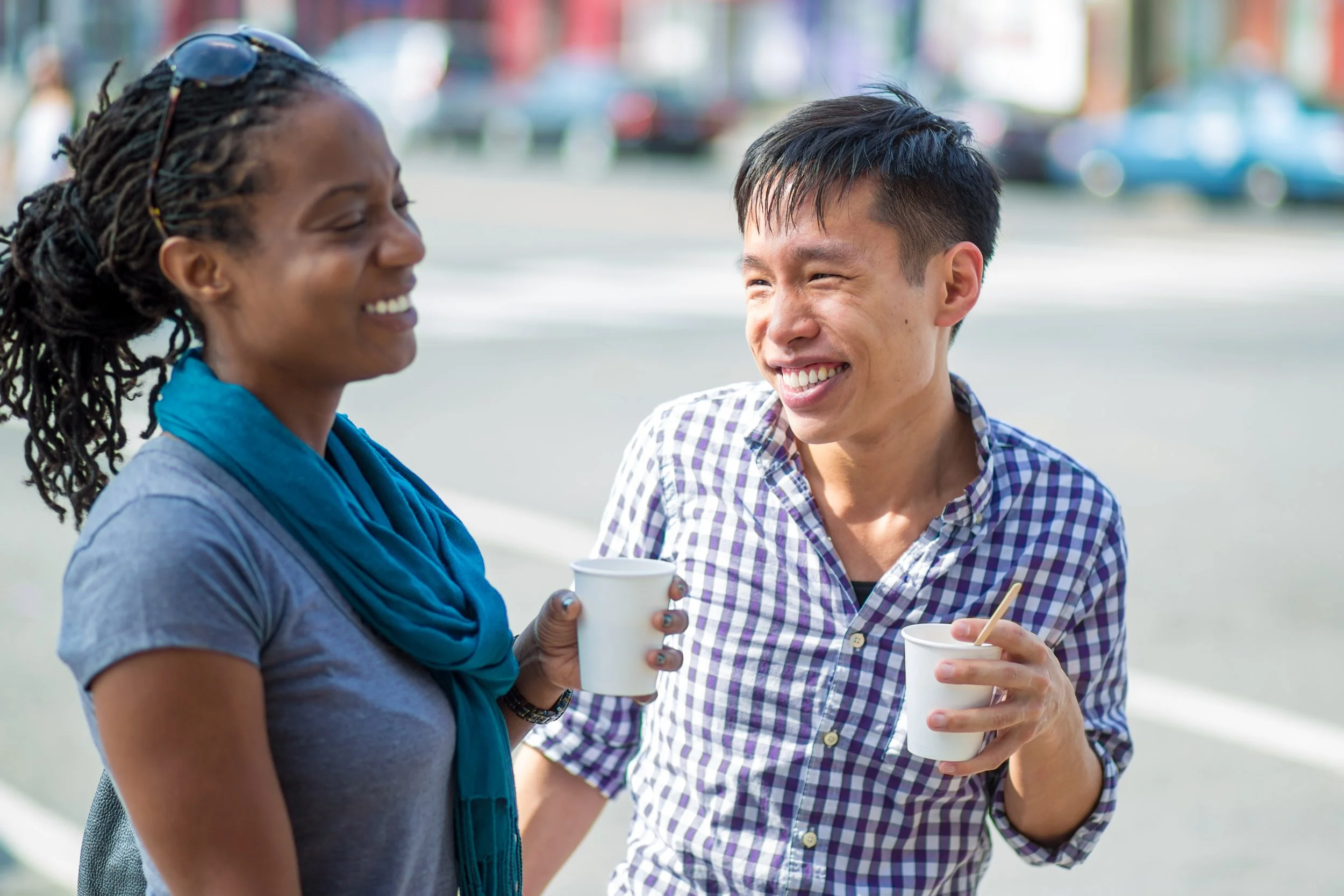 Two people talking and smiling outdoors, each holding a white cup.
