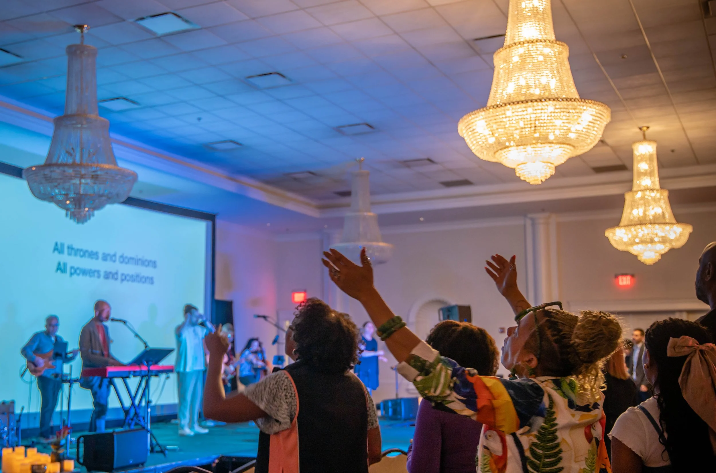 People attending church service and singing celebration, with a band performing on stage and a large screen displaying lyrics. Elegant chandeliers hang from the ceiling.