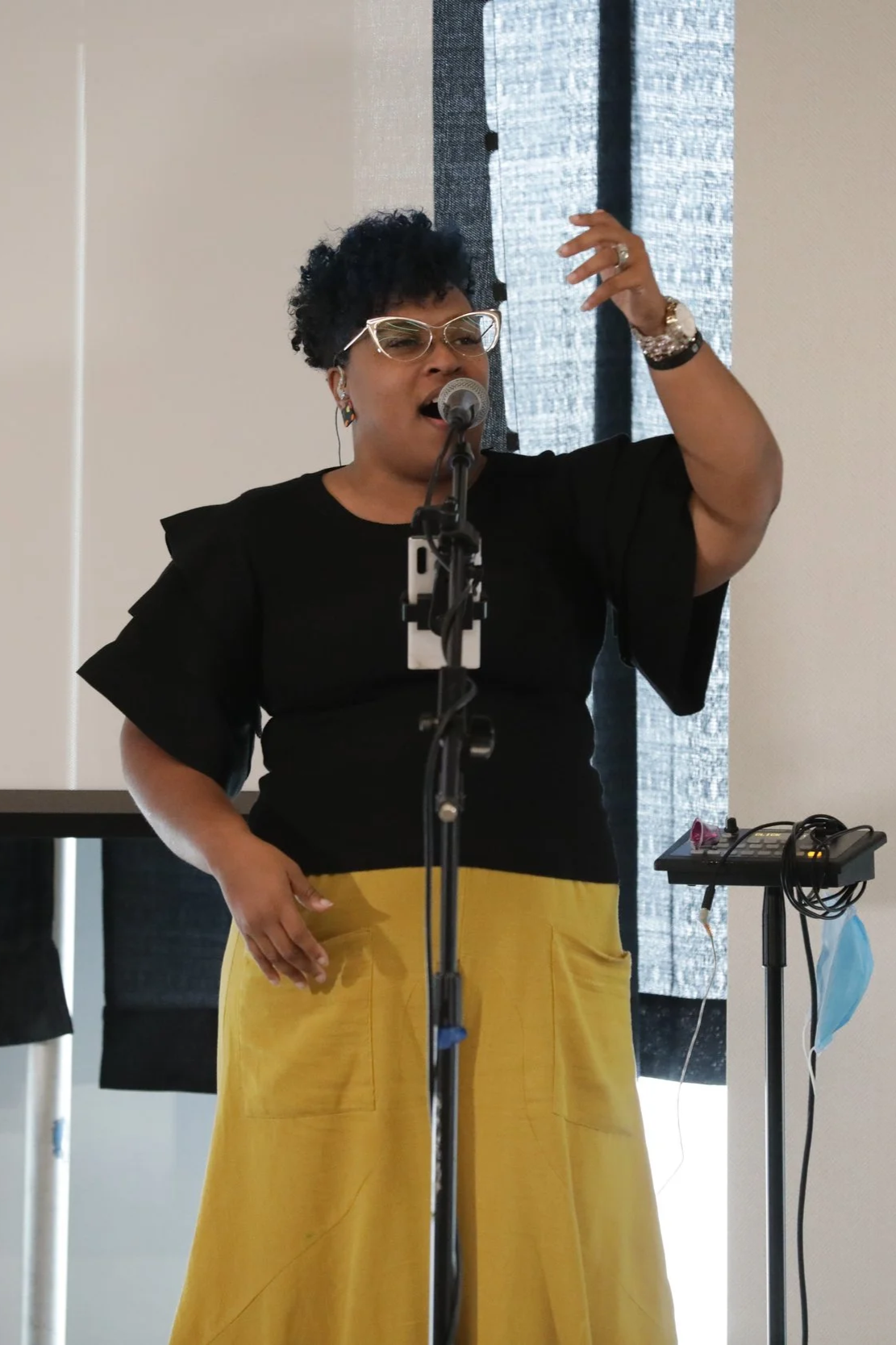 A woman singing into a microphone, wearing glasses, a black top with ruffle sleeves, and yellow pants. A person playing guitar is partially visible to the right.
