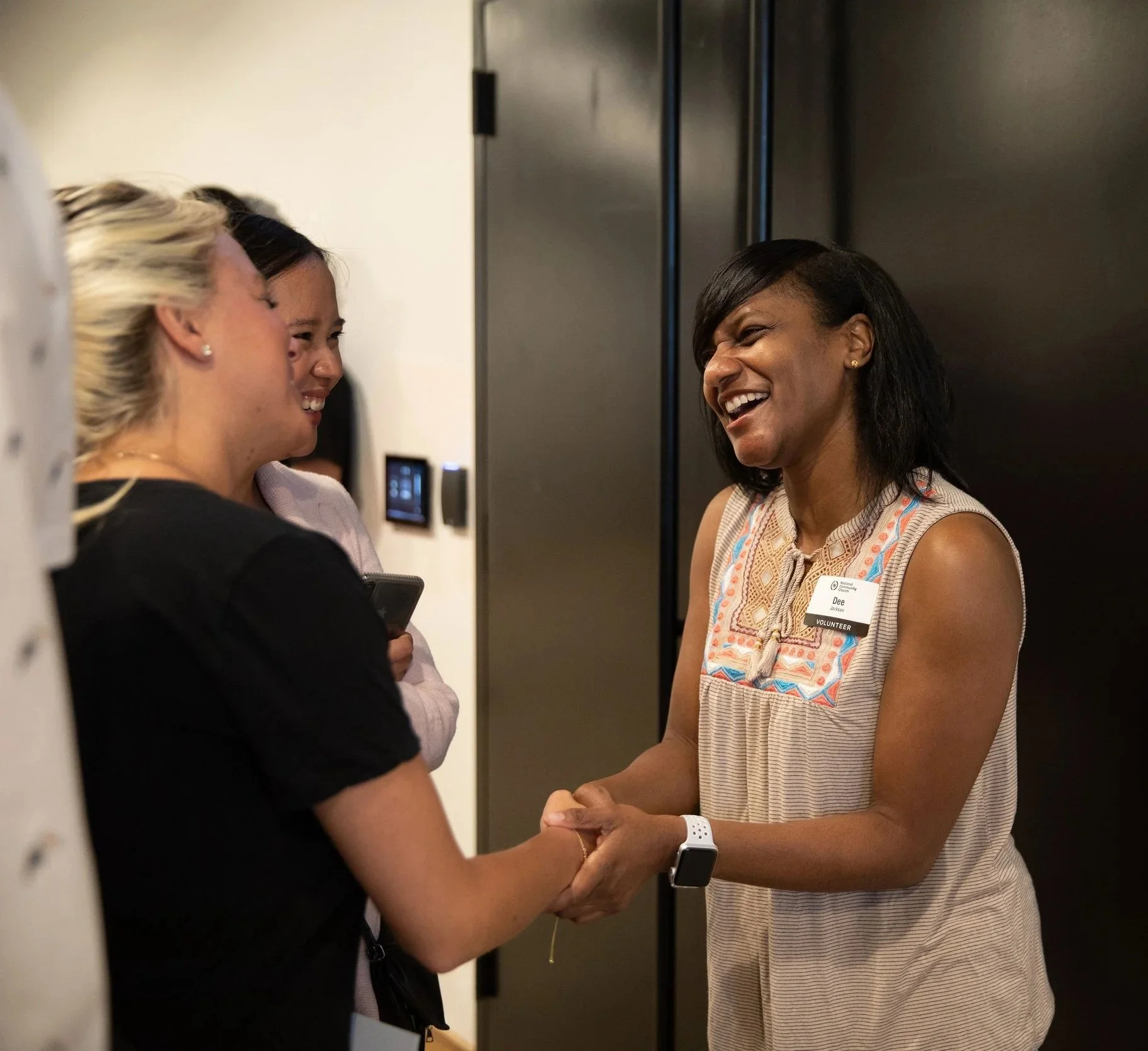 Two women shaking hands and smiling in a hallway near a door labeled 'VESTIBULE.' One woman is wearing a sleeveless top and a smartwatch; the other is holding a phone.