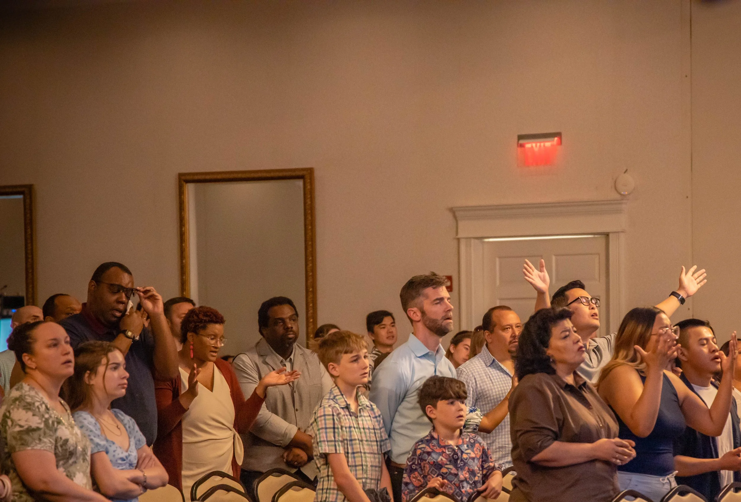 A diverse group of people in a church or conference hall, with some raising their hands and others with eyes closed in prayer or reflection.