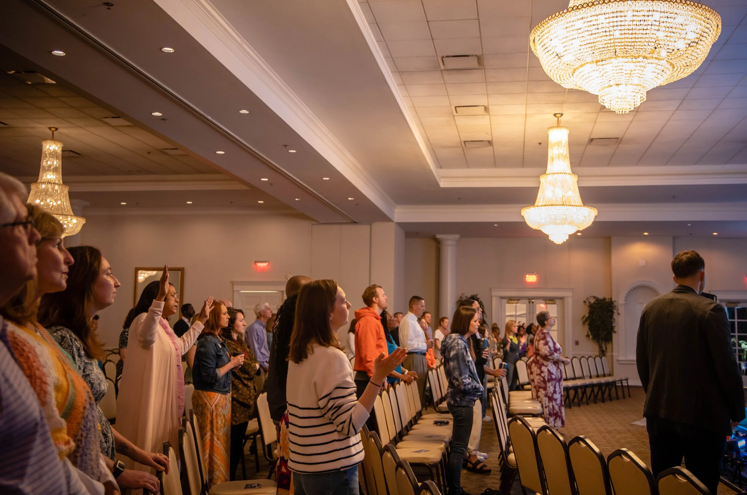 People standing with their hands raised in a large, well-lit conference or event room with chandeliers.