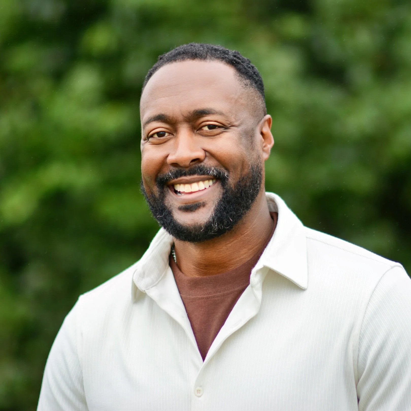 A smiling man with a white button up collared shirt, in front of greenery outdoors