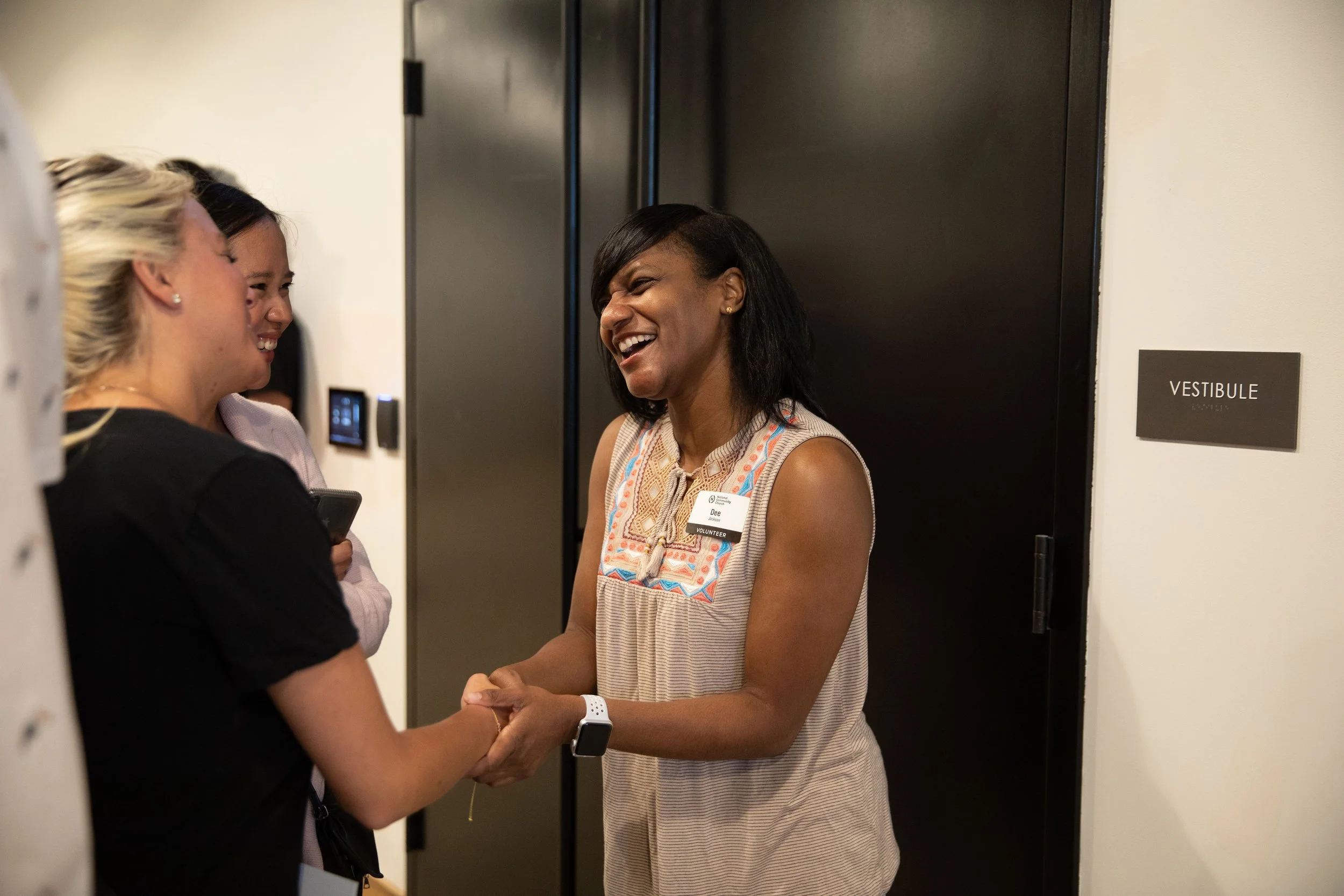 Two women shaking hands and smiling in a hallway near a door labeled 'VESTIBULE.' One woman is wearing a sleeveless top and a smartwatch; the other is holding a phone.